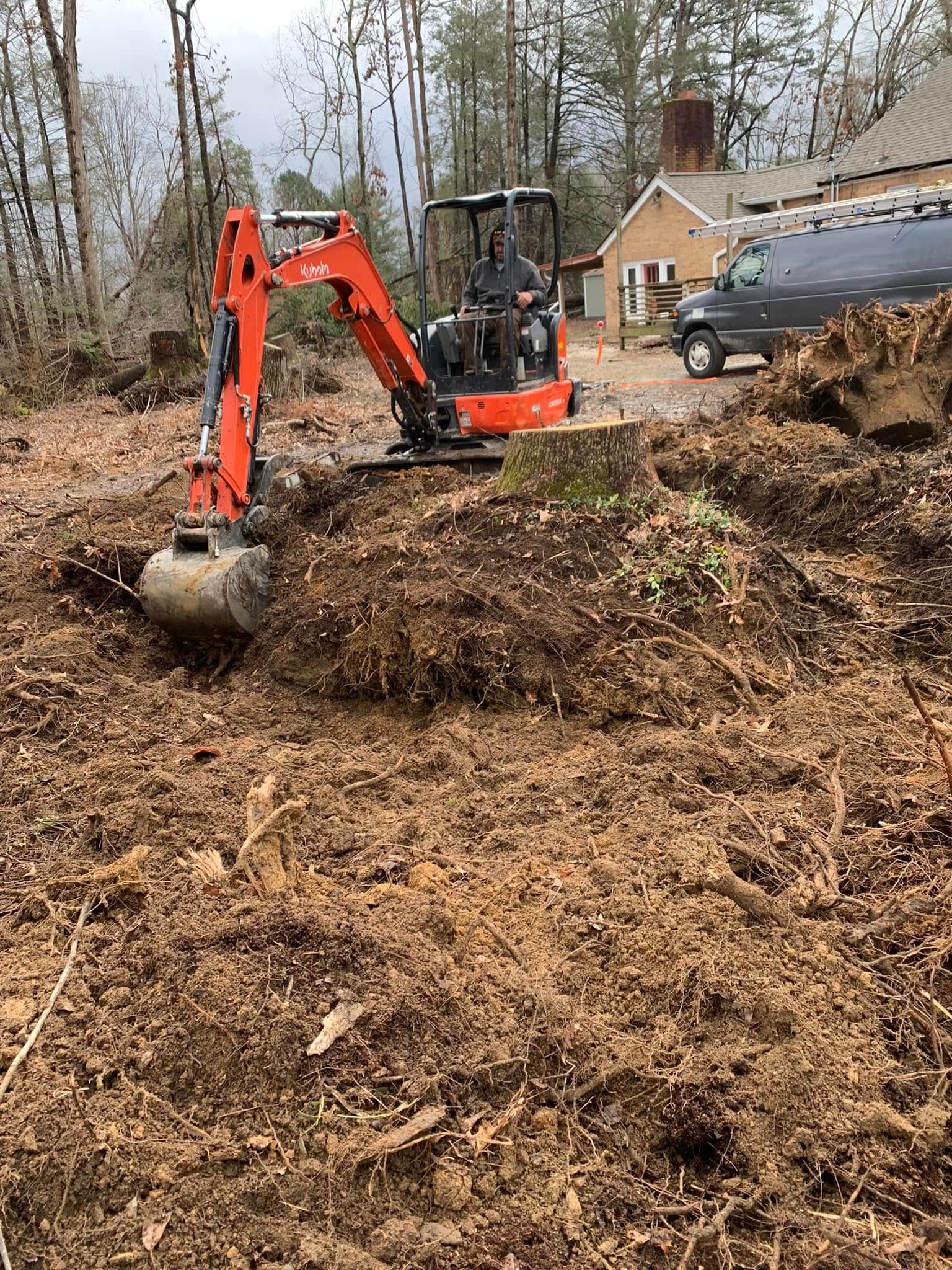 An orange mini excavator works on clearing earth and roots around a tree stump in a wooded yard near a house and van.