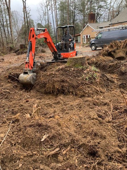 An orange compact excavator clears soil around a tree stump in a wooded yard near a house and a parked van.