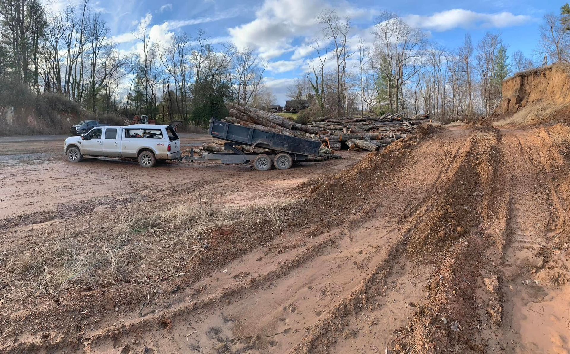 A white pickup truck with a trailer loaded with logs parked on a muddy, dirt-filled work site with trees in the background.