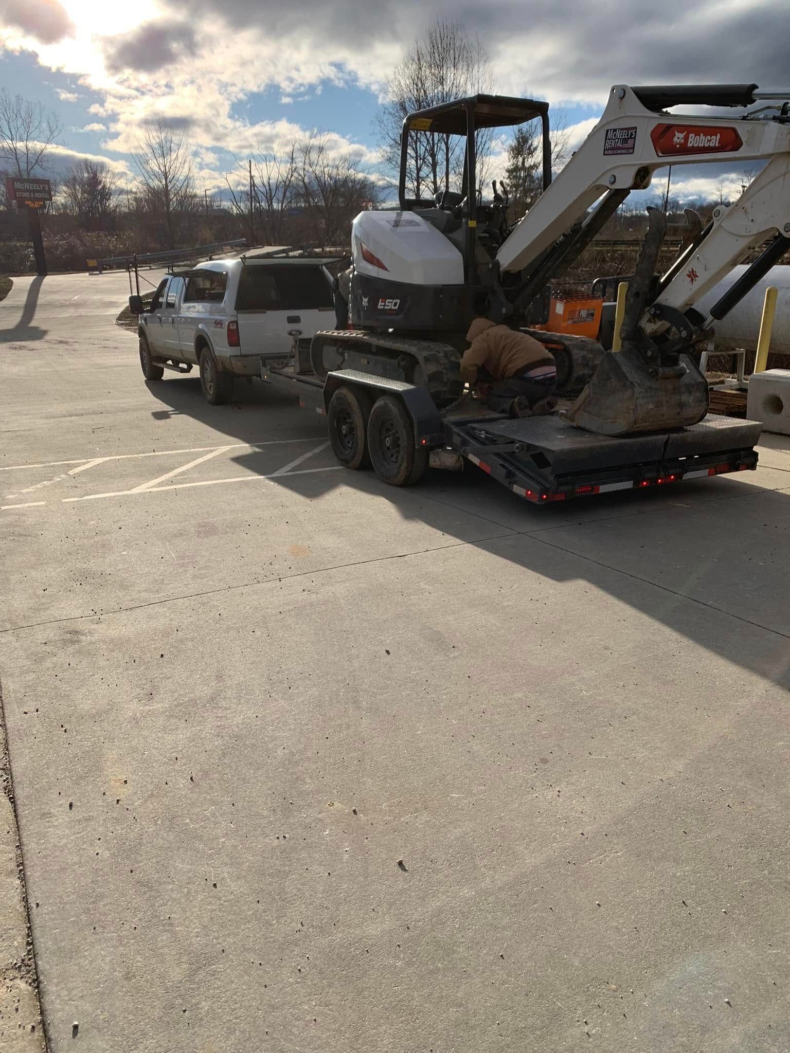 A white pickup truck hauls a flatbed trailer carrying a white and black compact excavator in a paved parking lot.