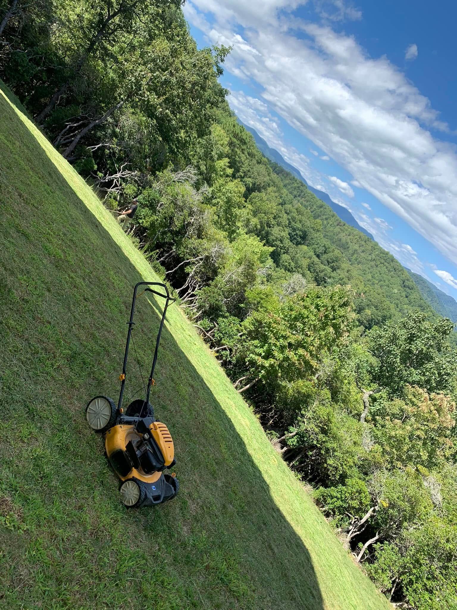 A yellow lawn mower sits on a green hillside slope overlooking a dense forest under a blue sky with white clouds.