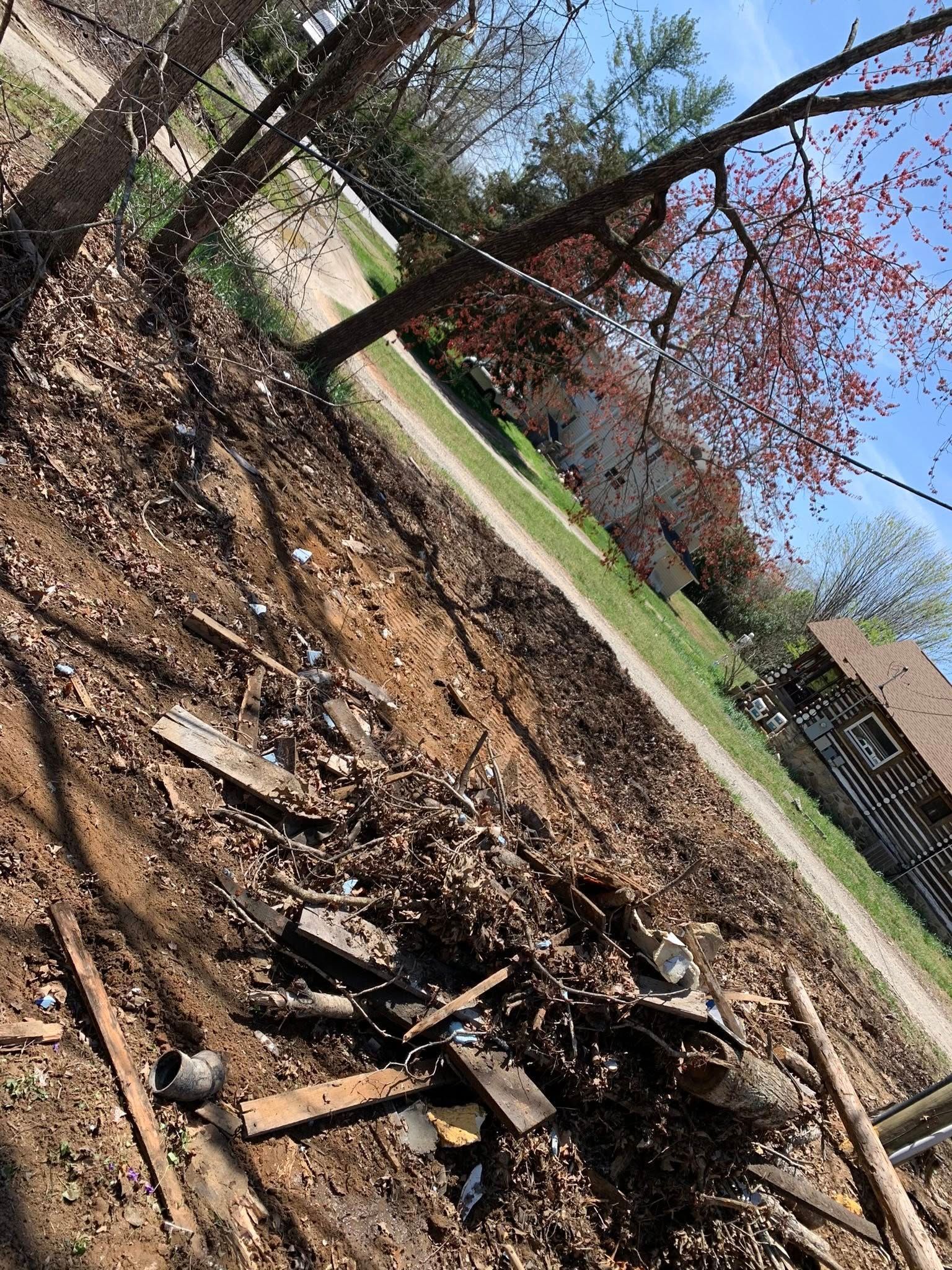 A pile of debris, including wooden boards and charred material, sits on a leaf-covered lawn near a house and trees.