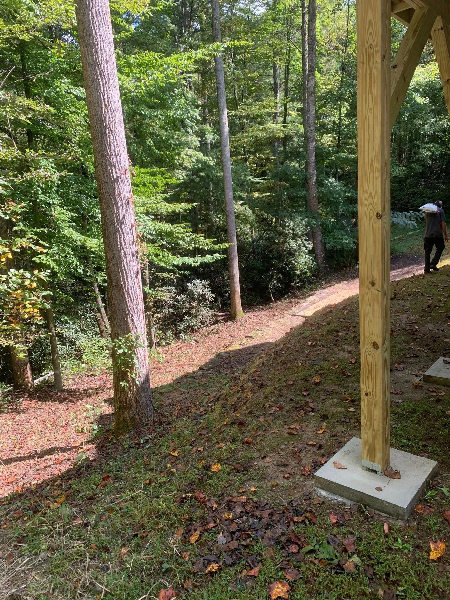 A wooden support post on a concrete base stands in a yard with a sloping hillside and dense forest in the background.