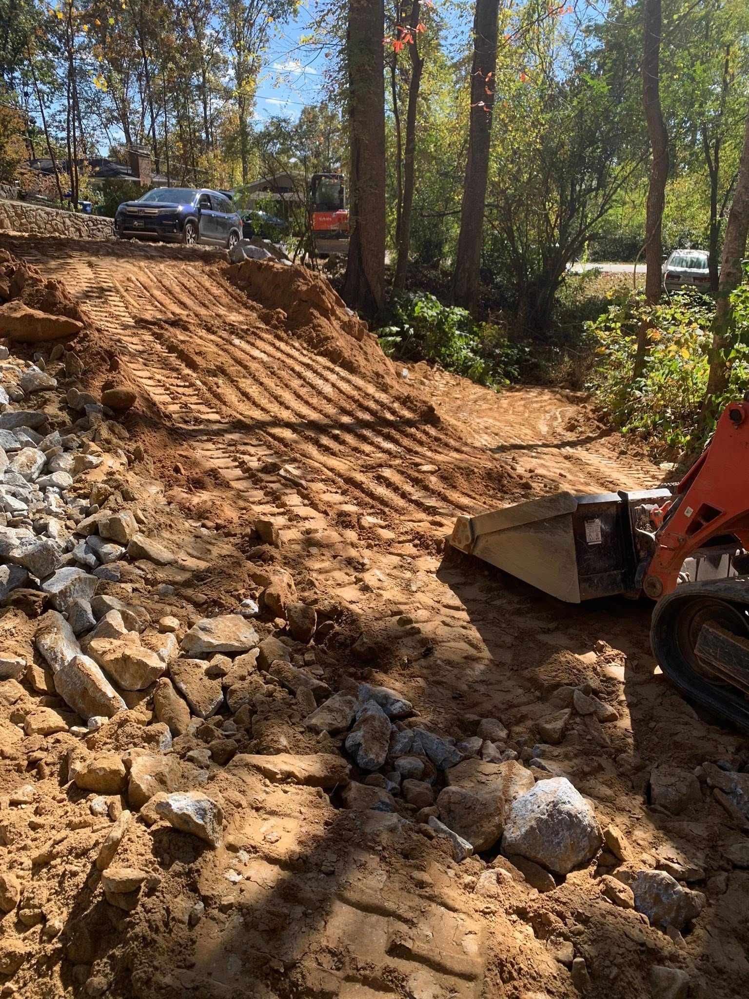 A compact excavator works on a dirt embankment, surrounded by trees with a car visible at the top of the slope.