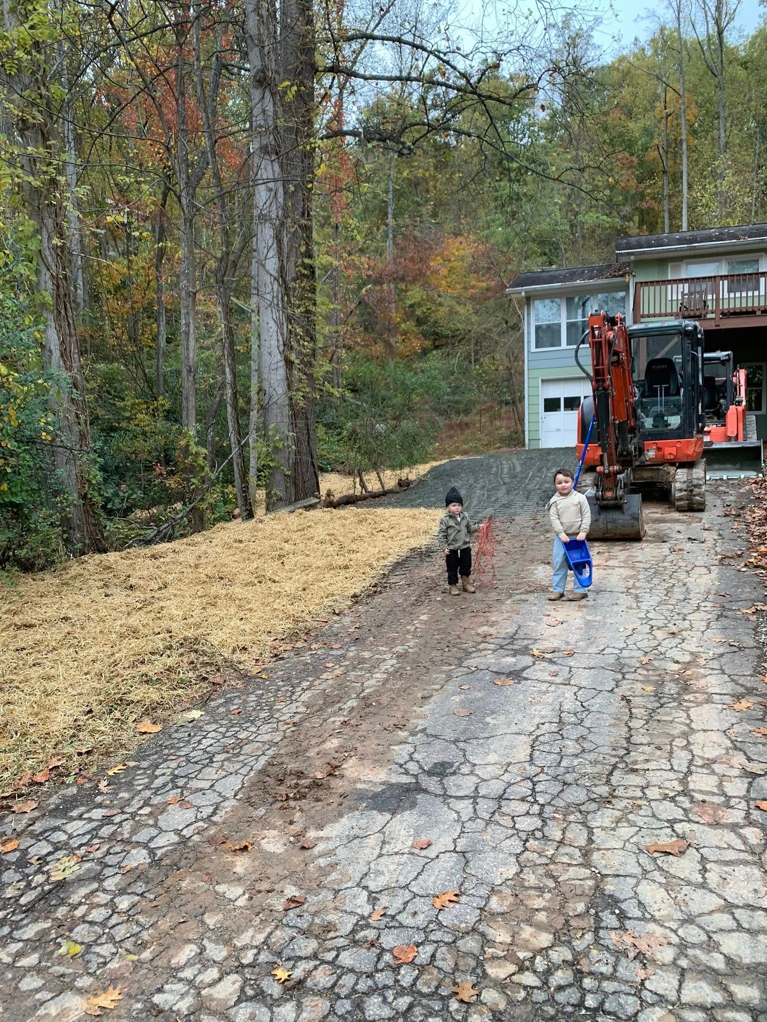 Two children stand on a stone driveway near an orange excavator and a house in a wooded area filled with autumn leaves.