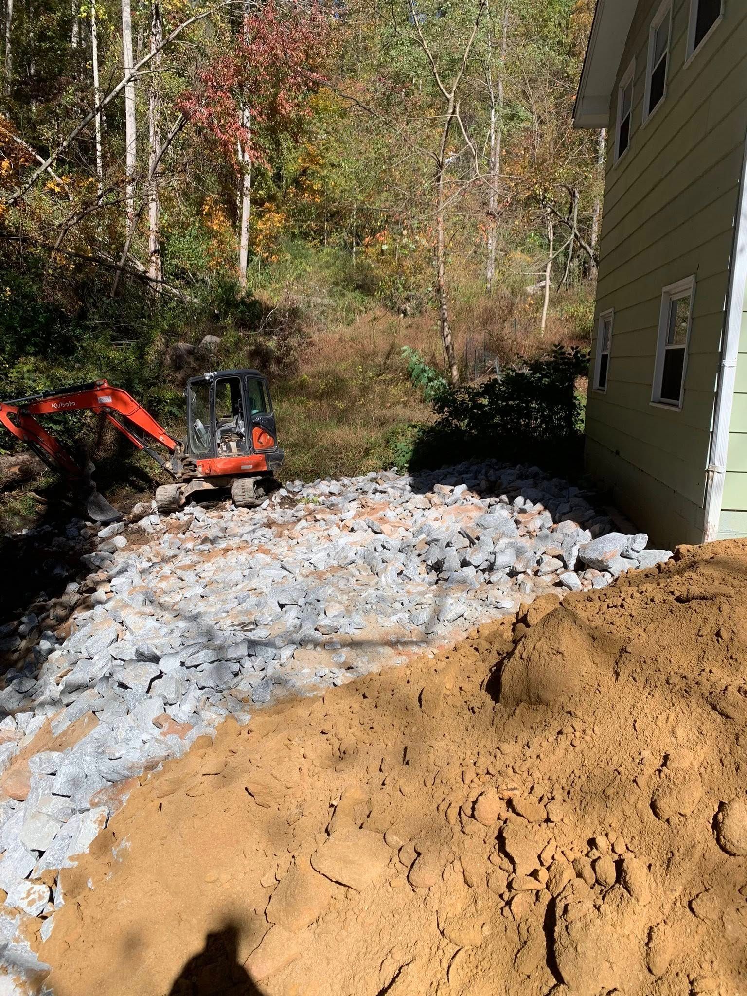 A small orange excavator sits near a house beside a pile of gravel and a large mound of dirt in a wooded area.