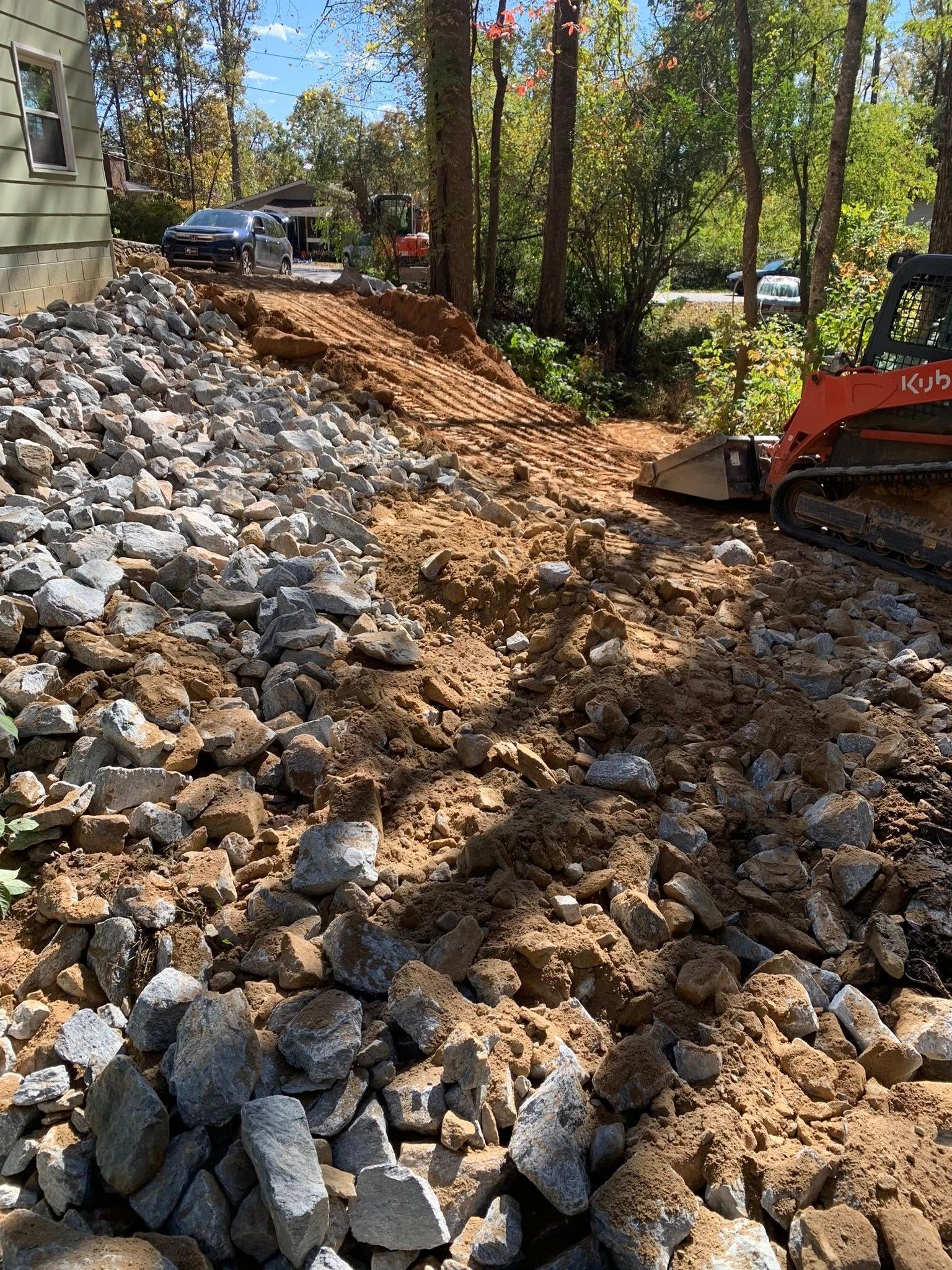 A small red excavator working on a dirt slope littered with large rocks next to a house.