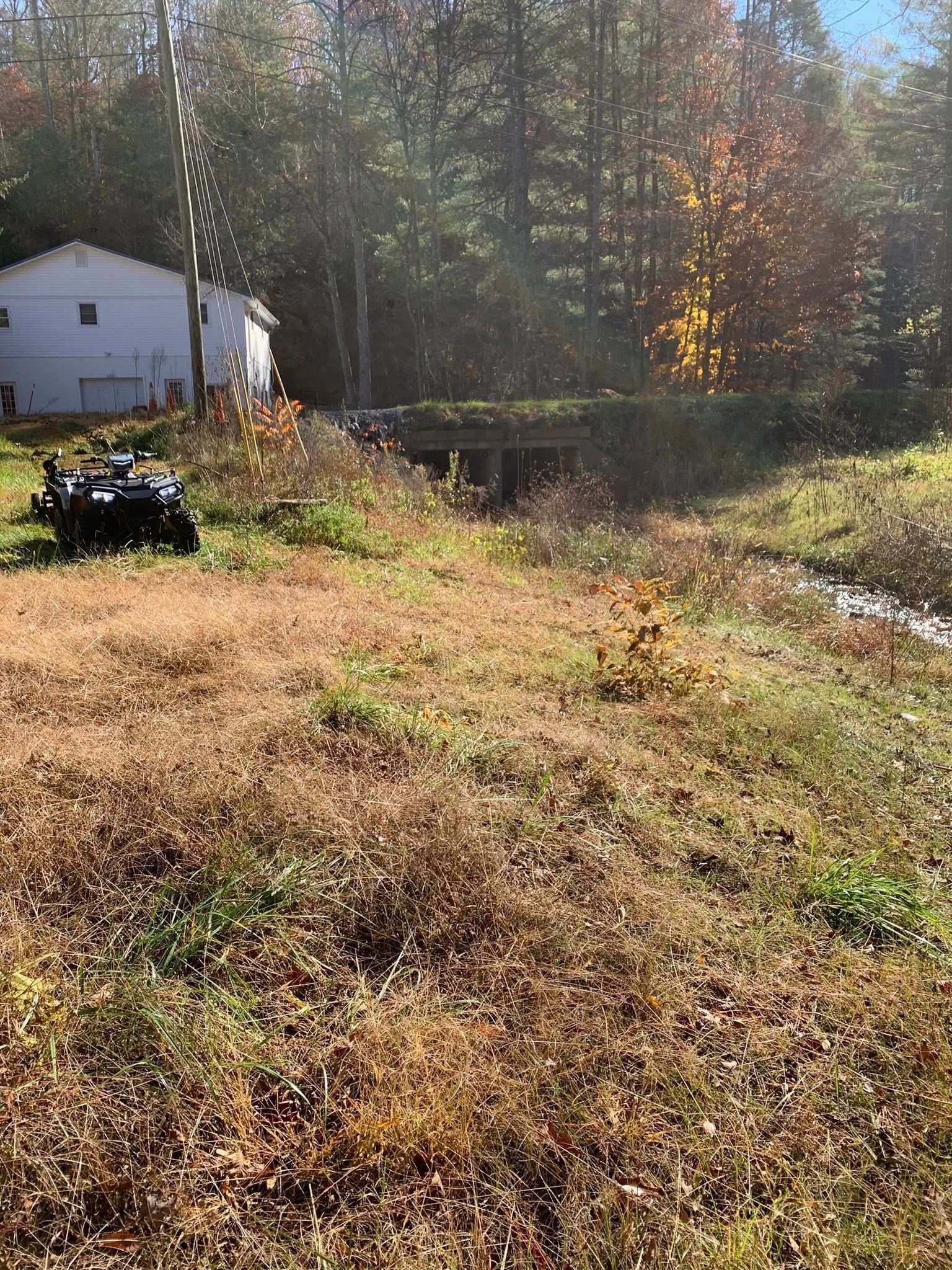 A white house sits near a wooded area with an ATV parked on a grassy slope next to a small stream.