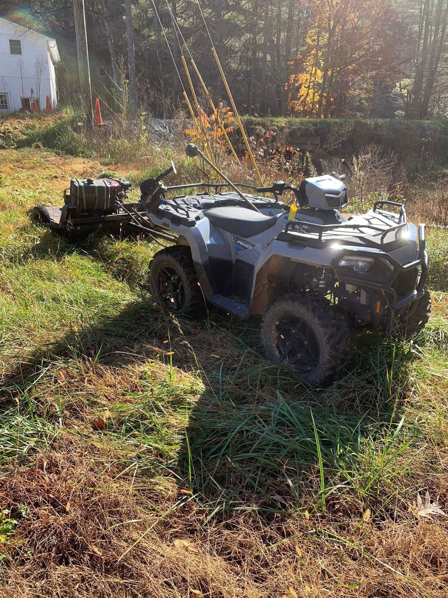 A grey ATV parked in a field, towing a small, dark tow-behind brush mower through tall, grassy terrain.