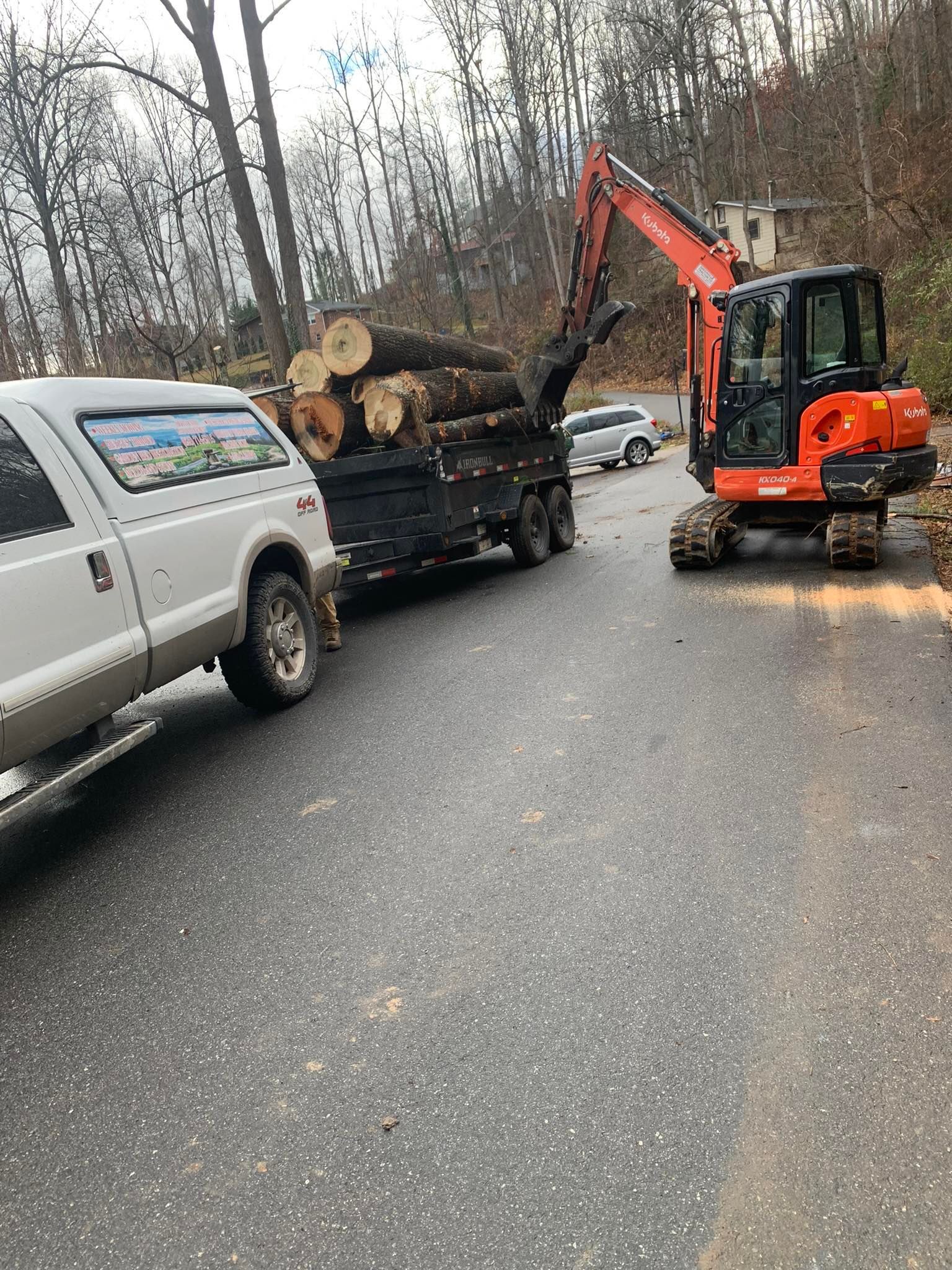 An orange excavator loading logs onto a trailer attached to a white pickup truck on a paved road.