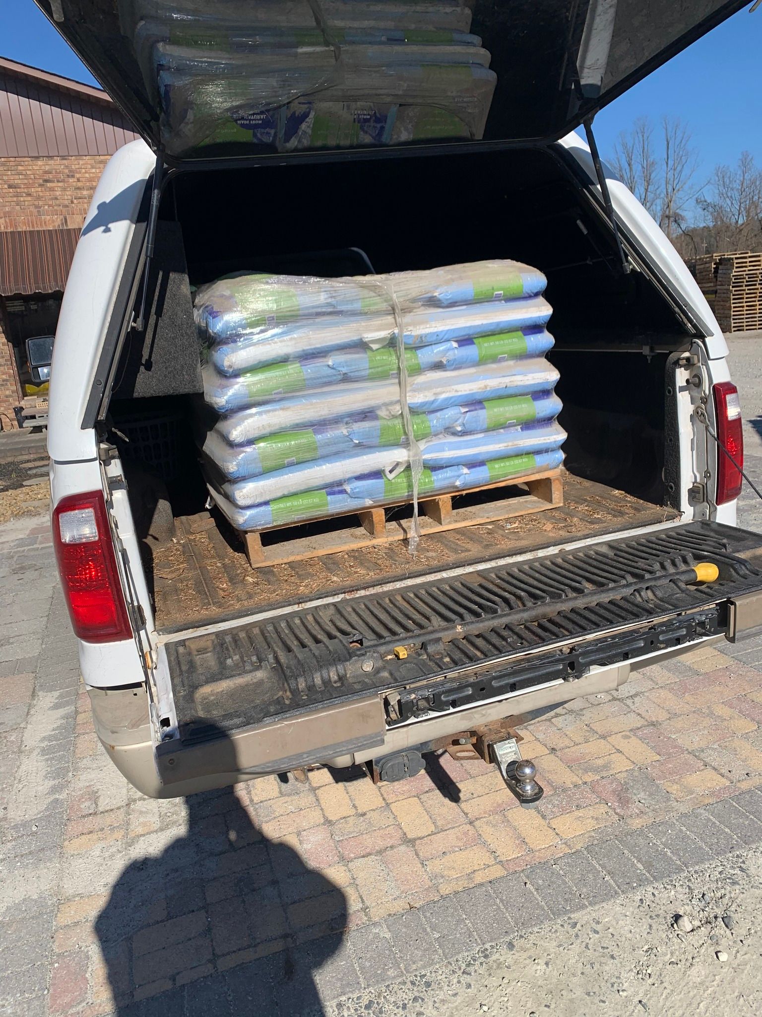 A wooden pallet stacked with blue and green bagged materials is loaded into the back of a white pickup truck.
