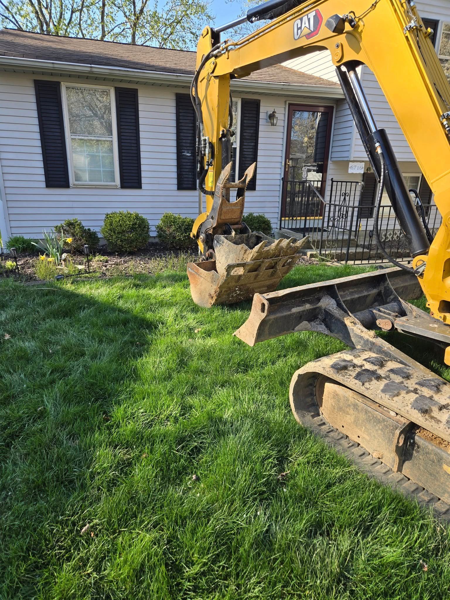 A yellow excavator is digging in the grass in front of a house.