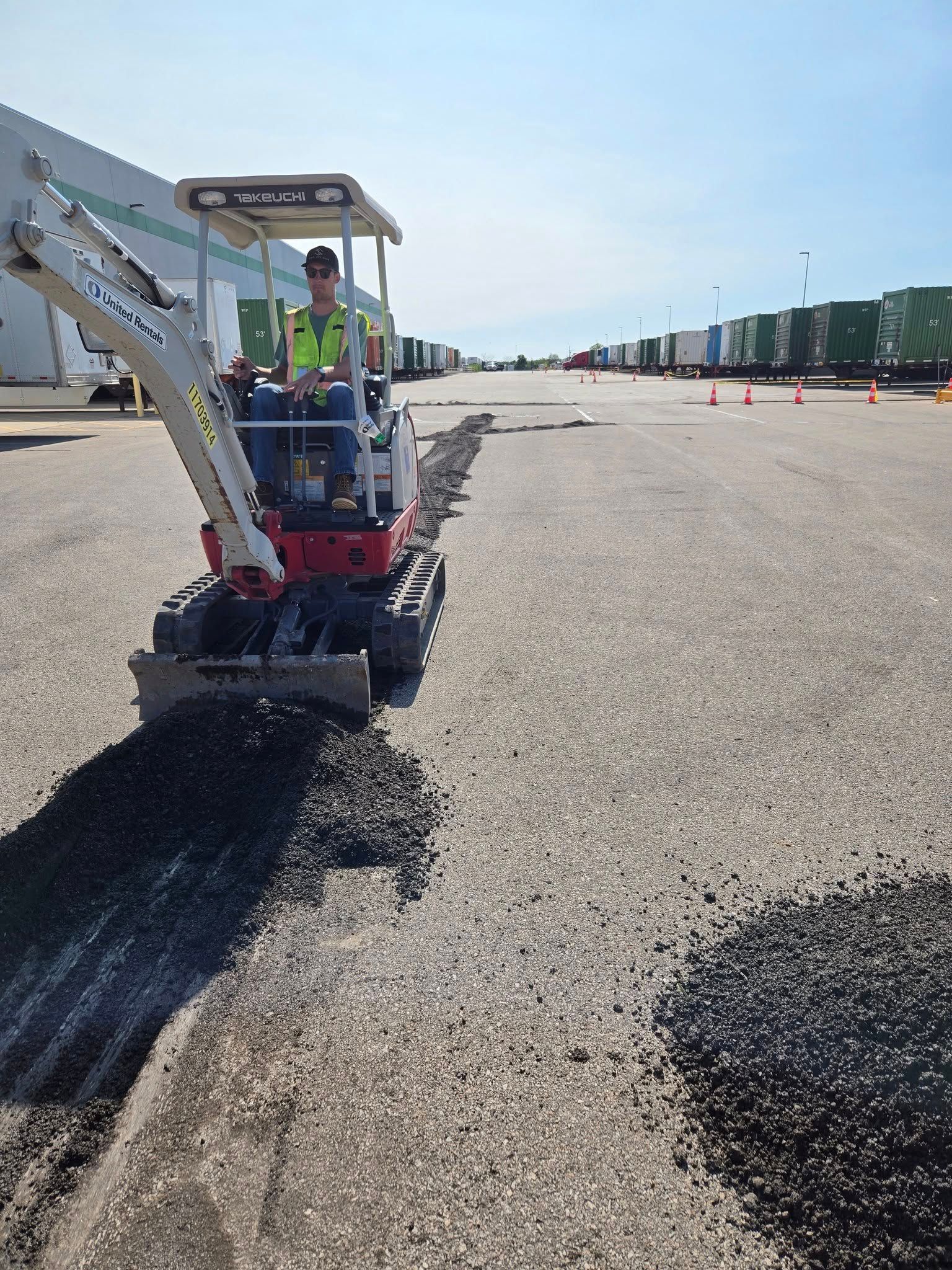 A man is driving a small excavator in a parking lot.