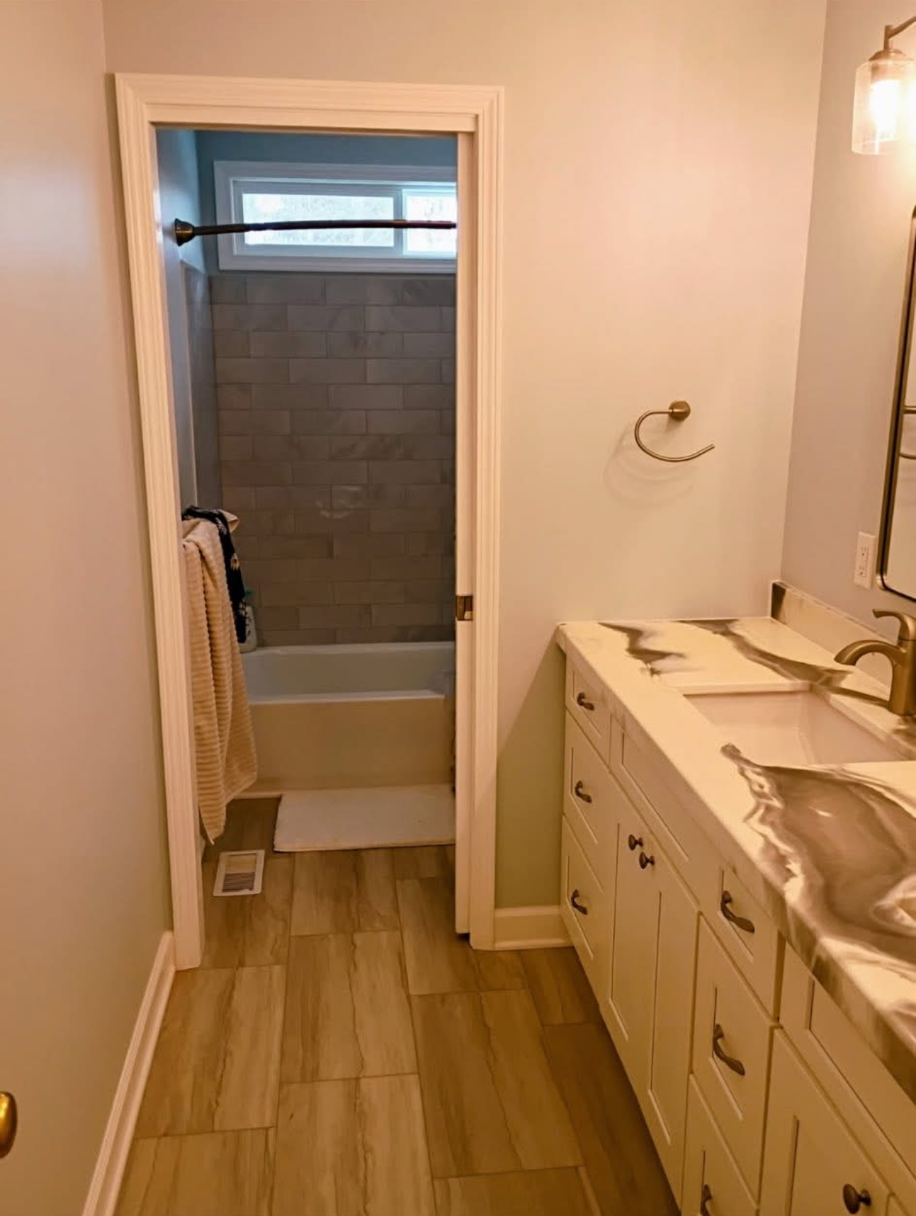 A view into a bathroom with wood-look tile flooring, a cream vanity with a marble countertop, and an adjacent shower room.