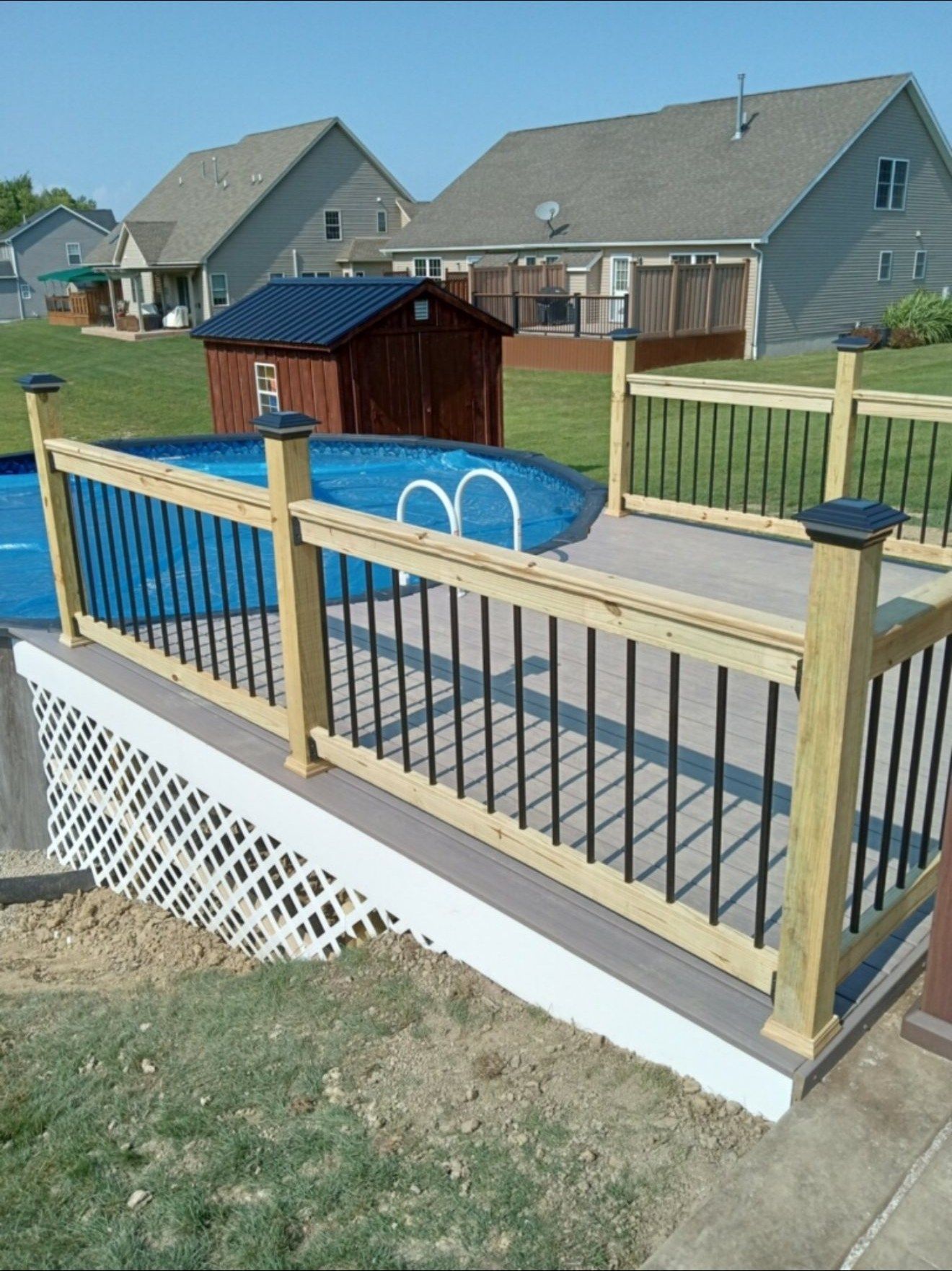 Wooden deck with black railing surrounds an above-ground pool in a backyard on a sunny day.