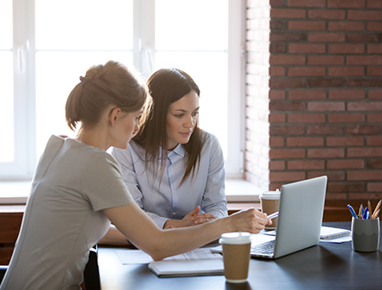 Two women are sitting at a table looking at a laptop computer.