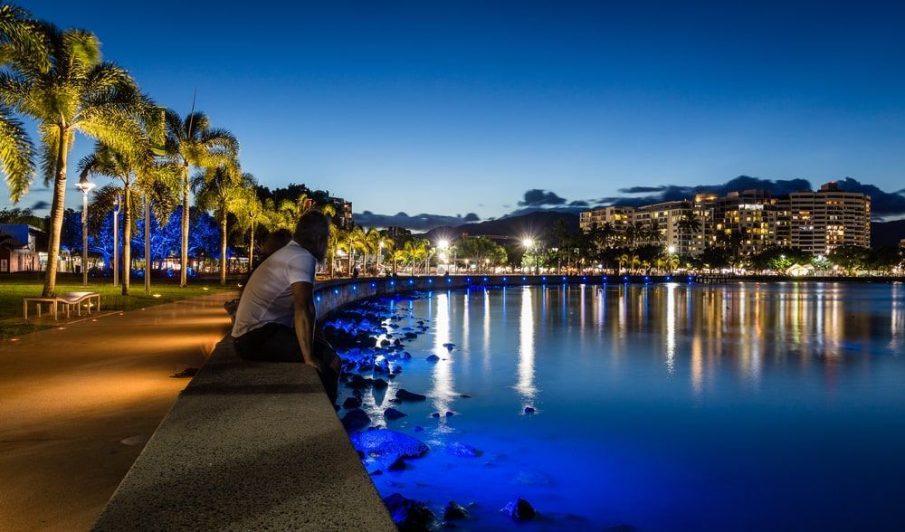 A Man Is Sitting On The Edge Of A Body Of Water At Night  — Lux Noir In  Cairns, QLD
