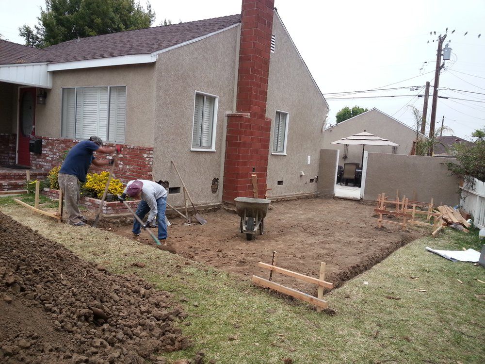 Two people digging in a yard near a house. One uses a shovel, the other works with a level. Brown earth is piled up.