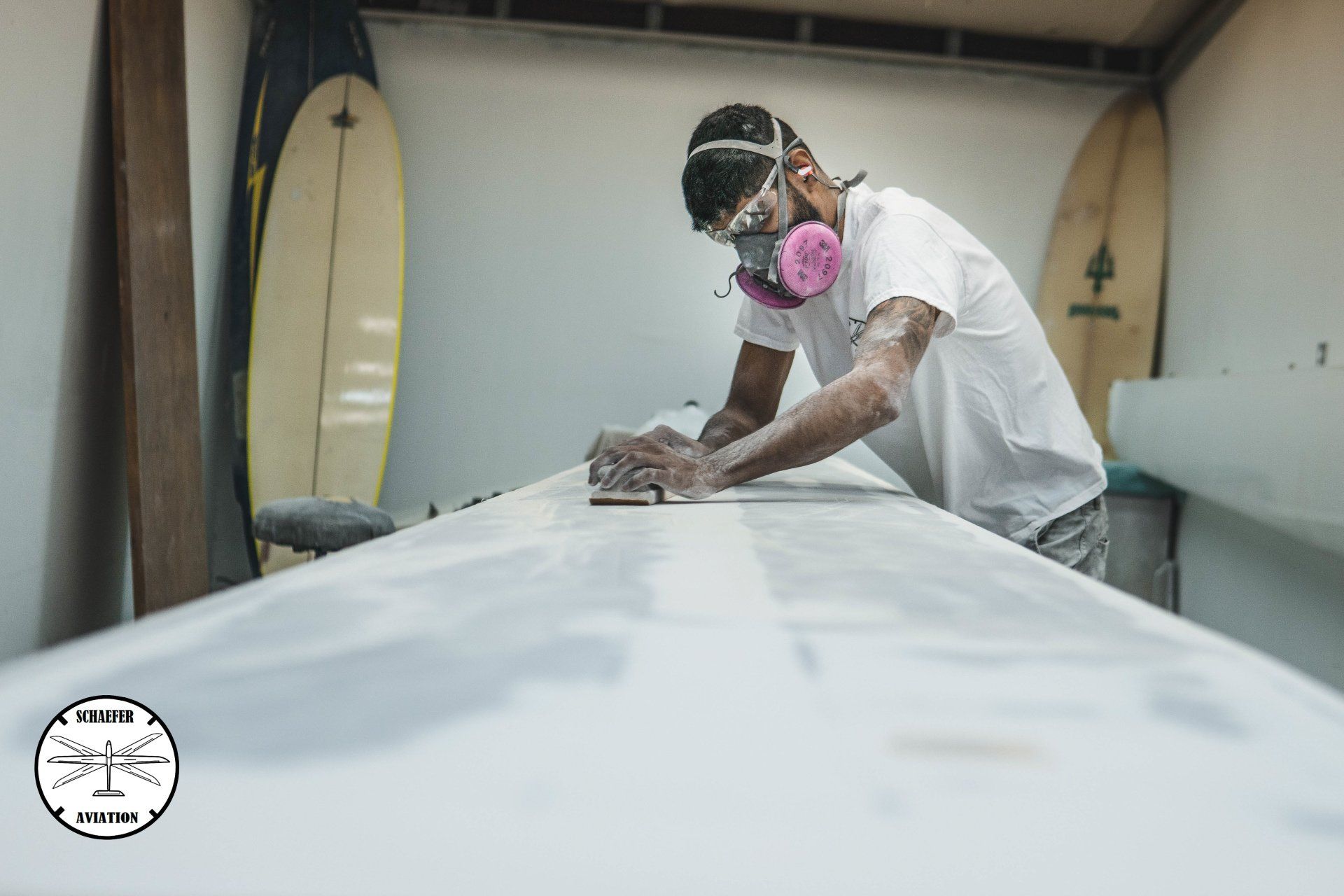 A man wearing a mask is sanding a surfboard in a garage.