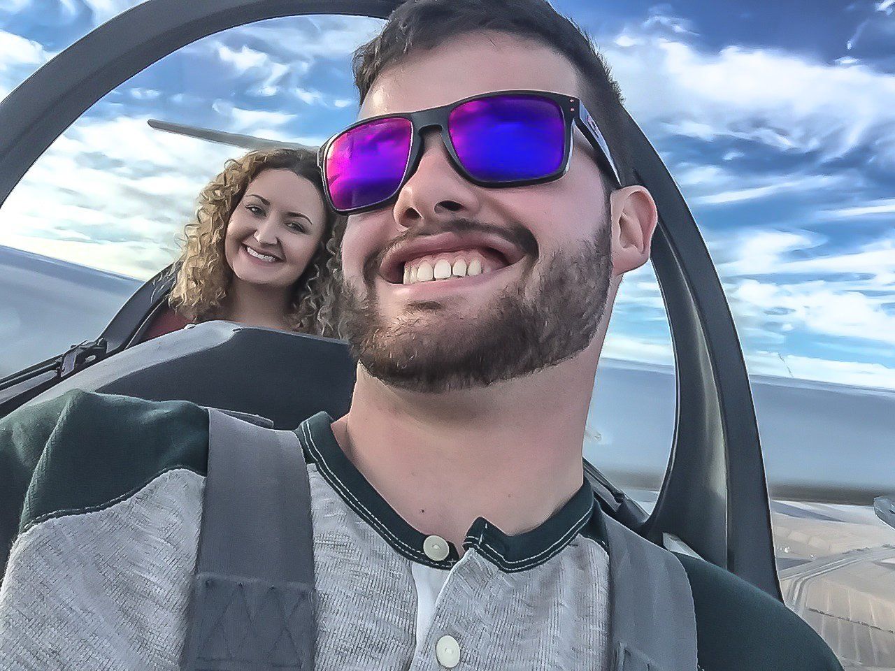 A man wearing sunglasses is smiling in a plane with a woman behind him