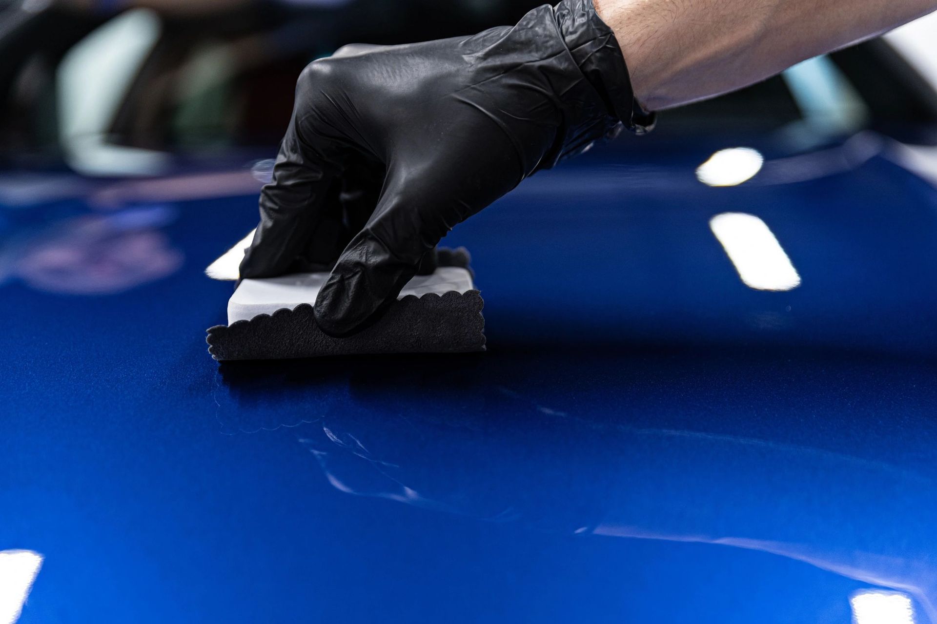 A person wearing black gloves is polishing a blue car with a sponge.