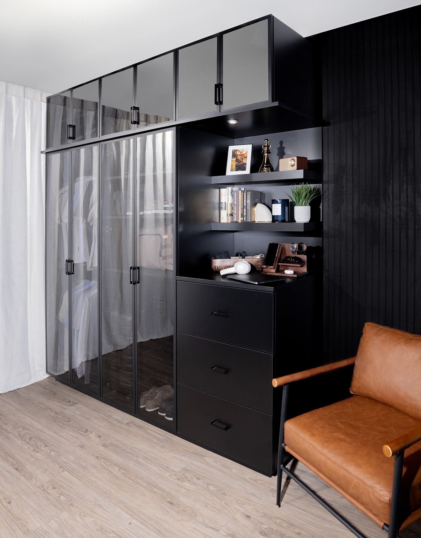Modern bedroom corner with black wardrobes, glass doors, shelving, and a tan chair on light wood flooring