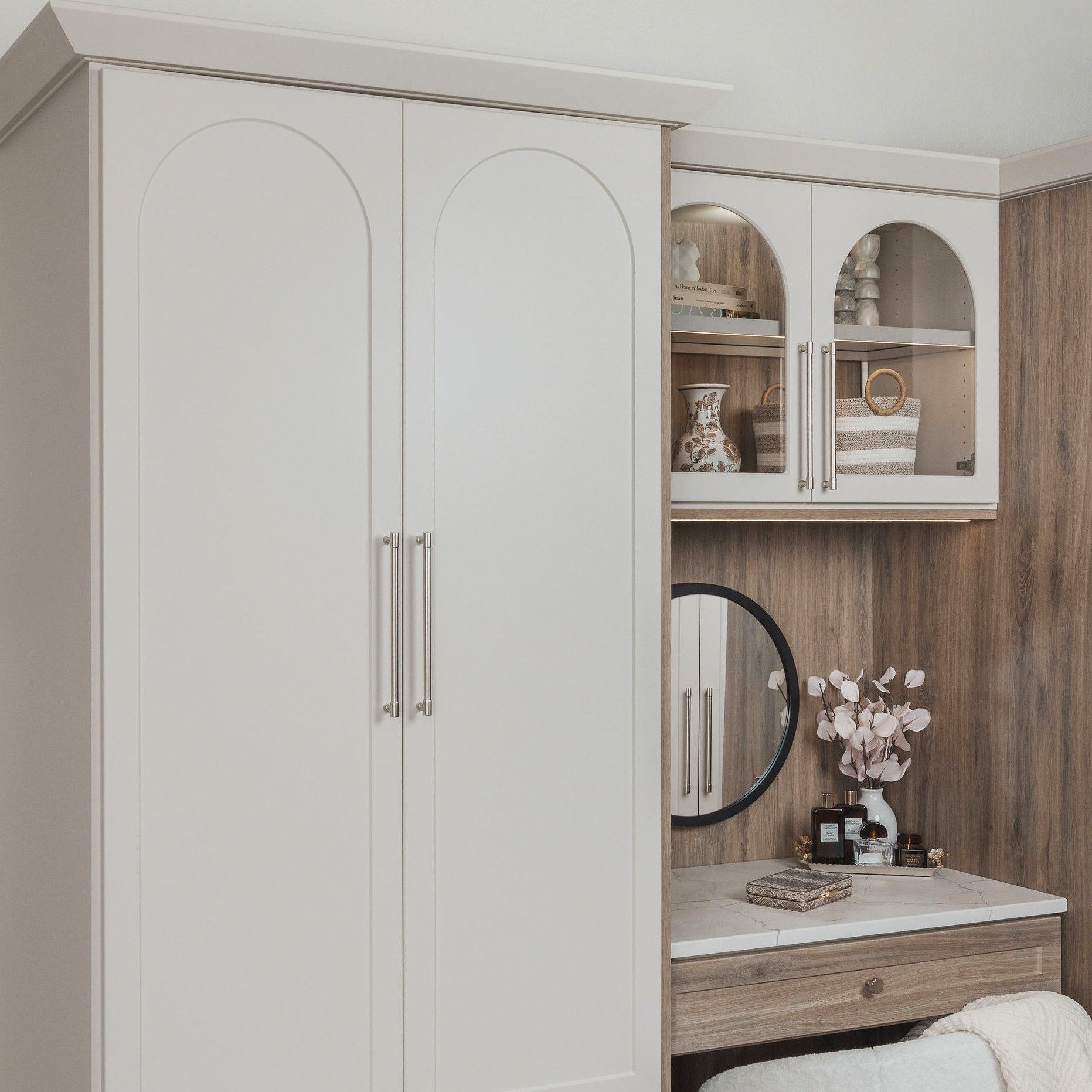 White arched wardrobe beside a wooden vanity with mirror, shelf decor, and flowers in a cozy bedroom corner