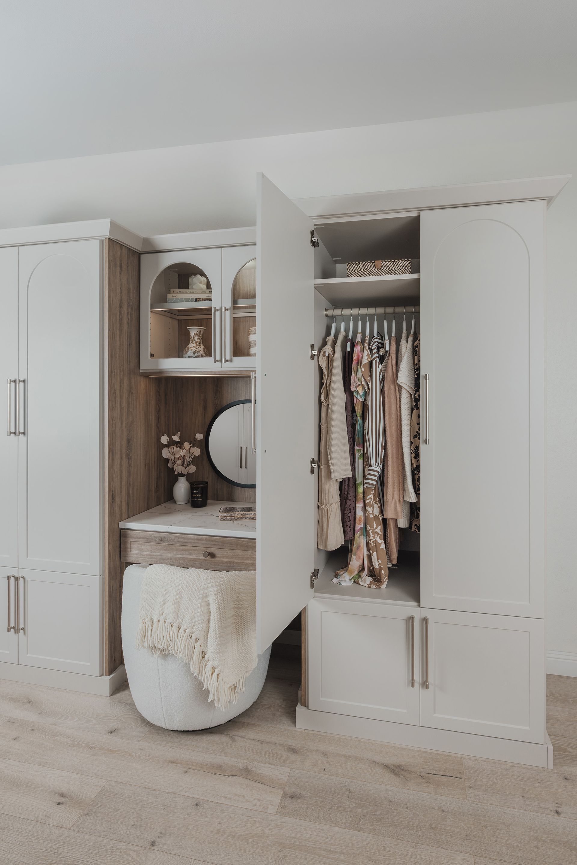 Open closet with shelves, hanging clothes, mirror, and woven ottoman in a bright bedroom corner.