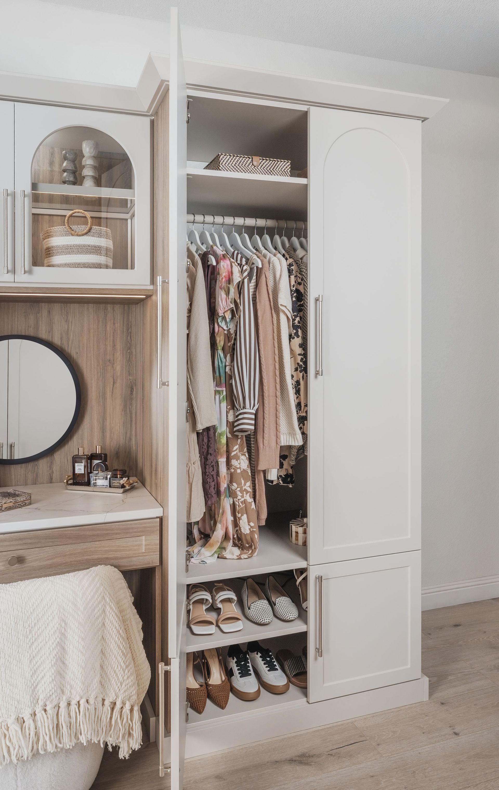 White open wardrobe with hanging clothes, shelves, and shoes in a bedroom beside a vanity.