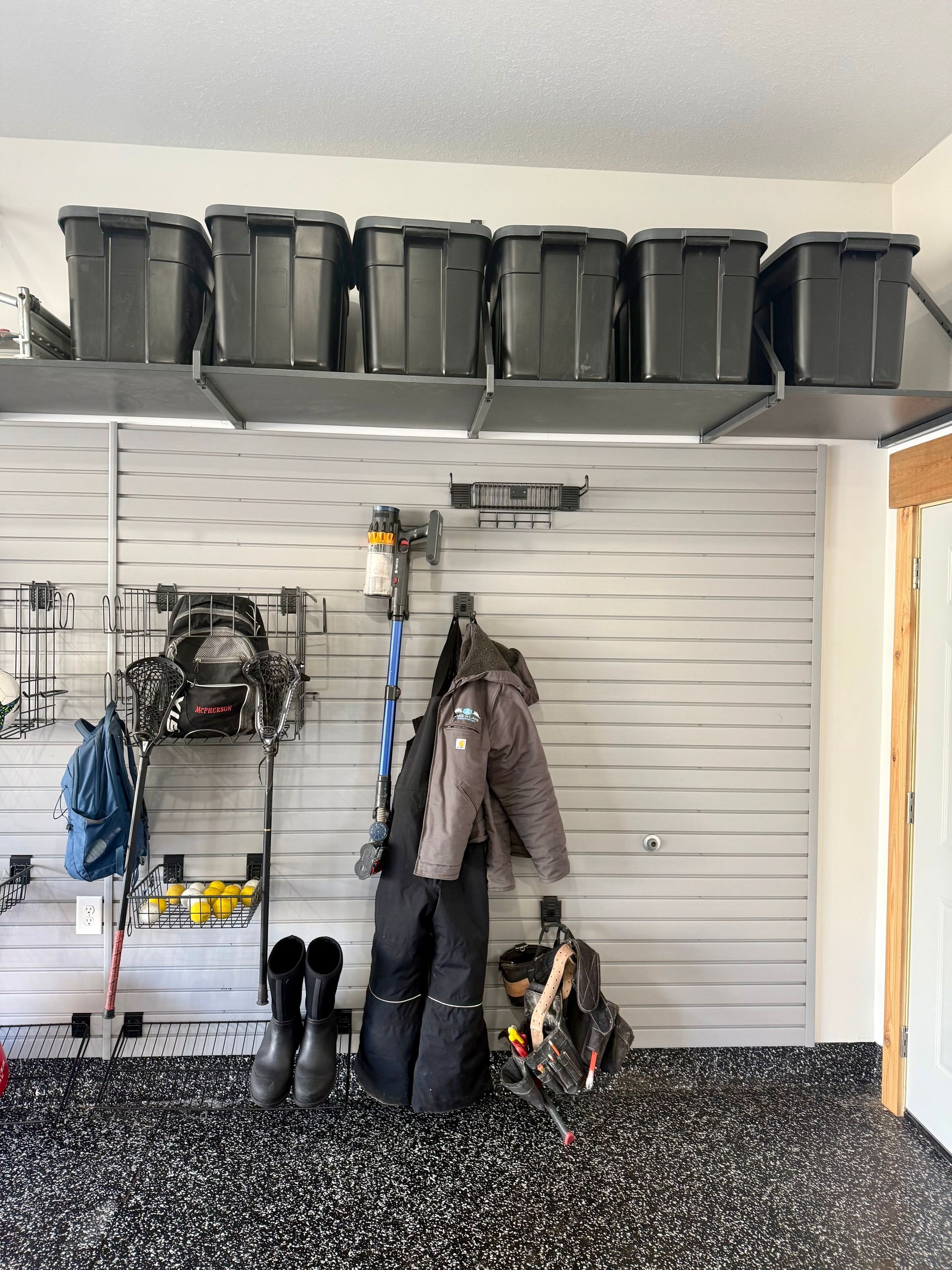 Garage wall with slatwall storage, hanging tools, coats, boots, and black bins on overhead shelves.