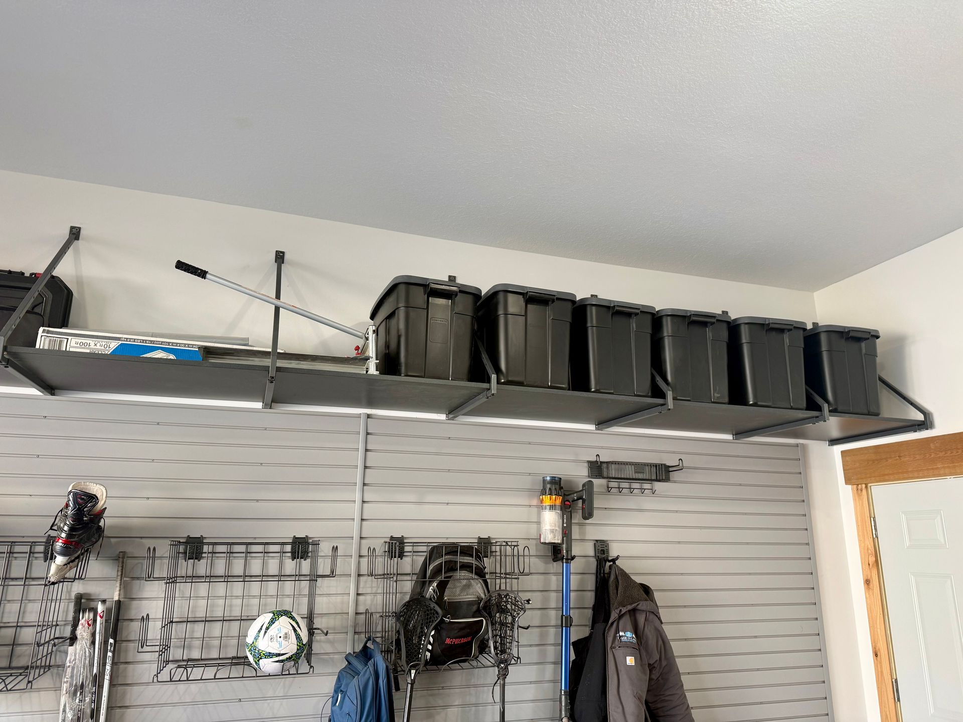 Garage wall with a metal shelf holding black storage bins, hanging tools, and jackets on a slat wall.