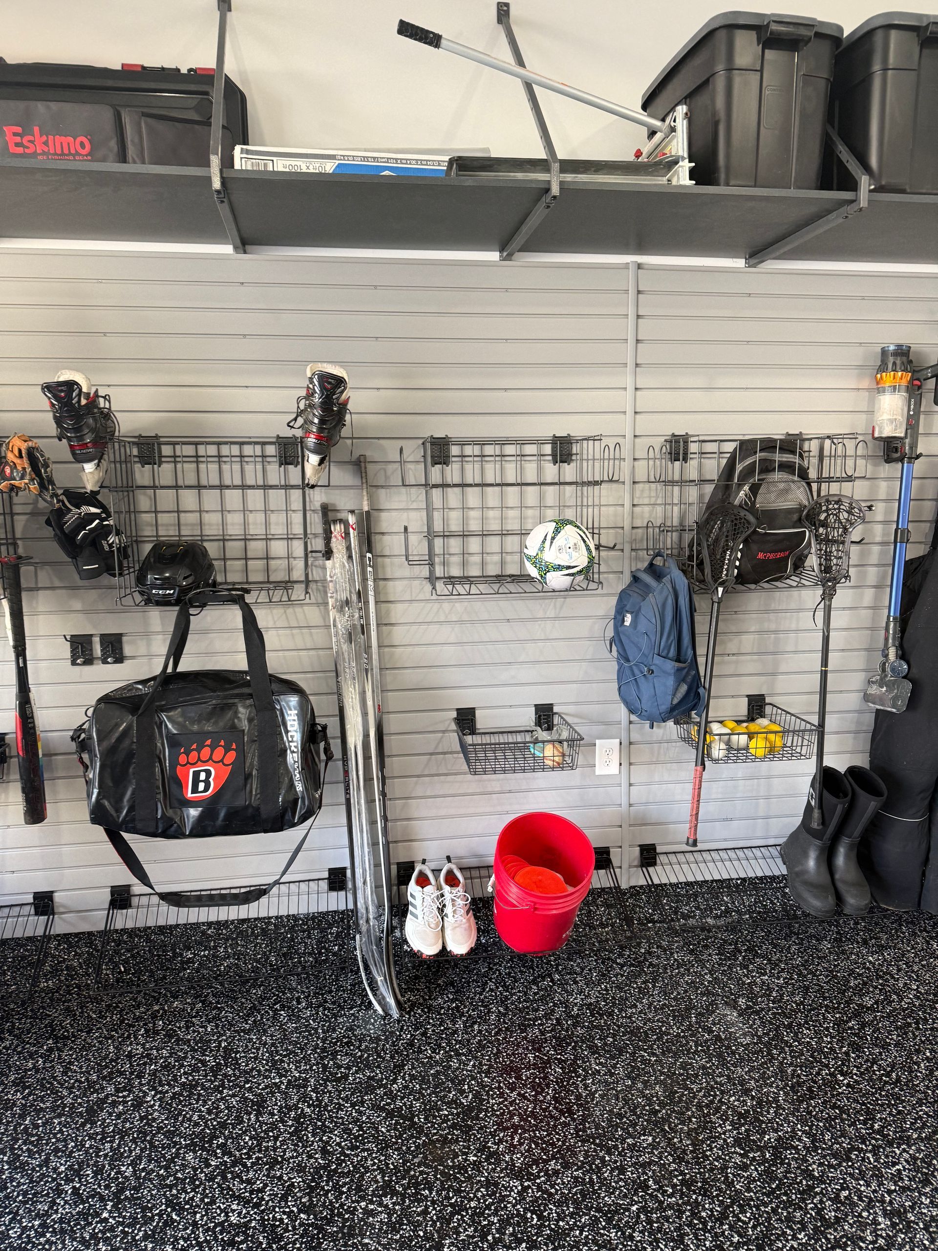 Garage wall with fishing rods, gear bags, boots, and a red bucket on speckled floor.