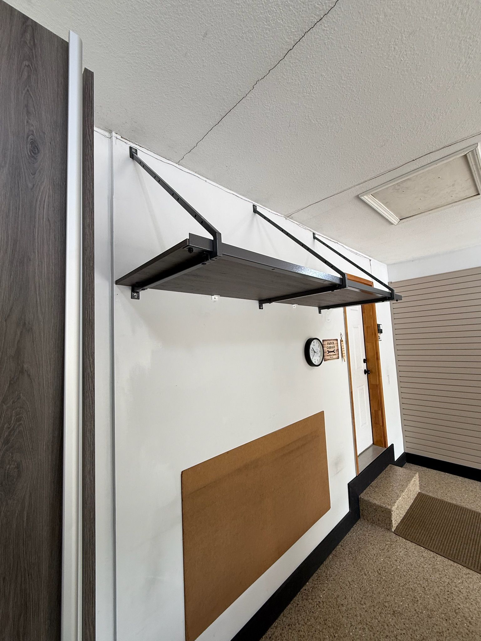 Wall-mounted folding shelf or seat in a hallway, with a white wall and carpeted floor.