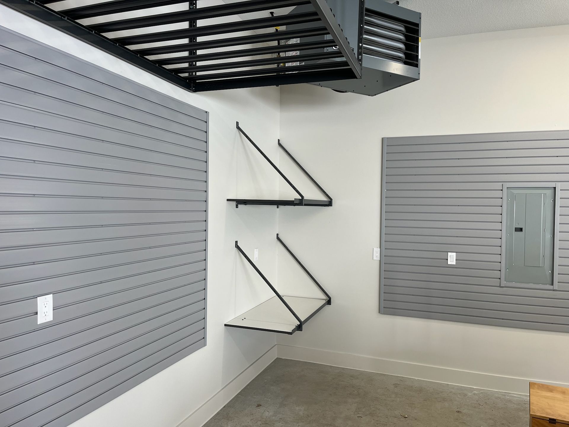 Empty industrial room with gray slat walls, two folding shelves, and a black overhead mezzanine.