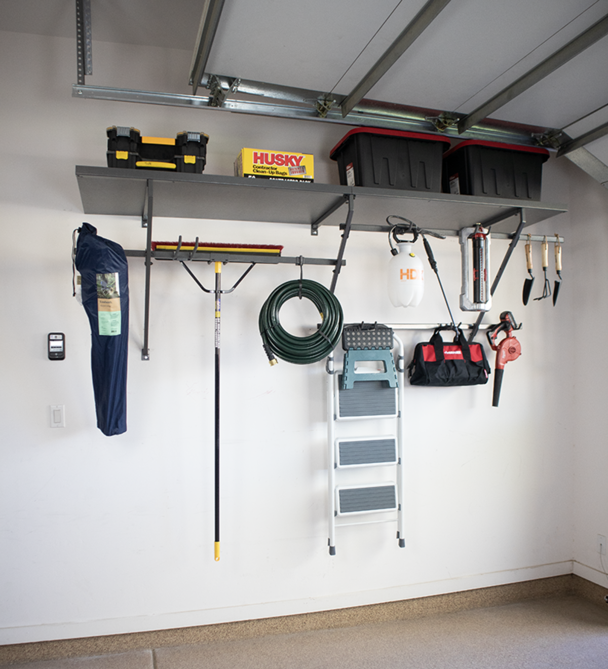 Garage wall with shelves and stored tools, including a ladder, hose, and toolboxes.