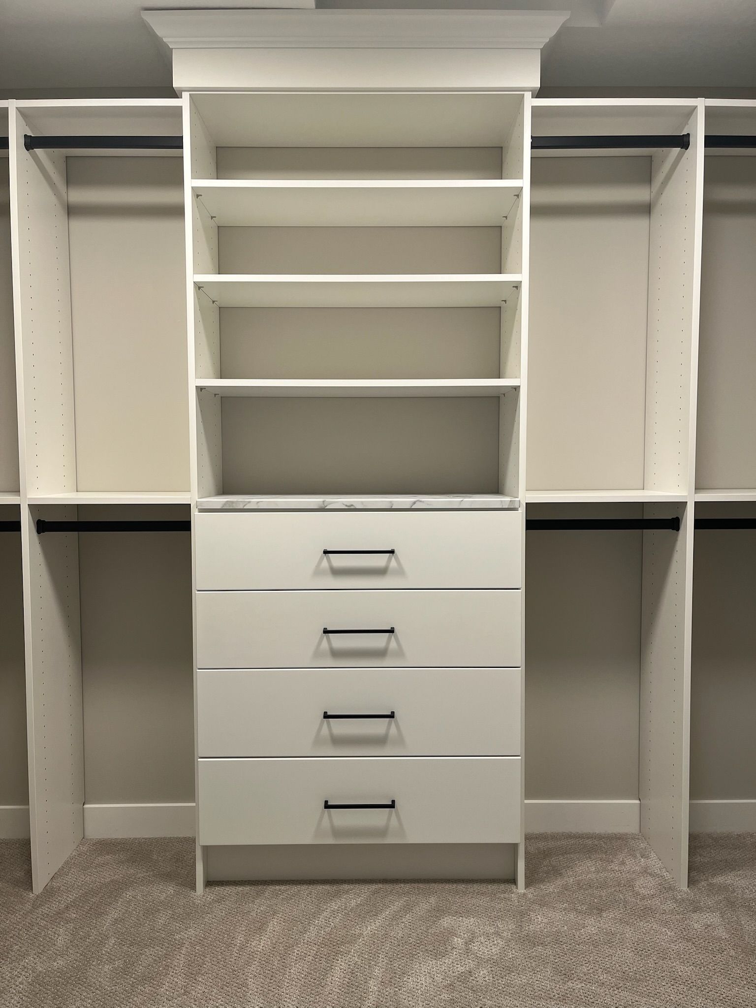 White built-in closet organizer with shelves and four drawers, centered between hanging spaces on carpet.