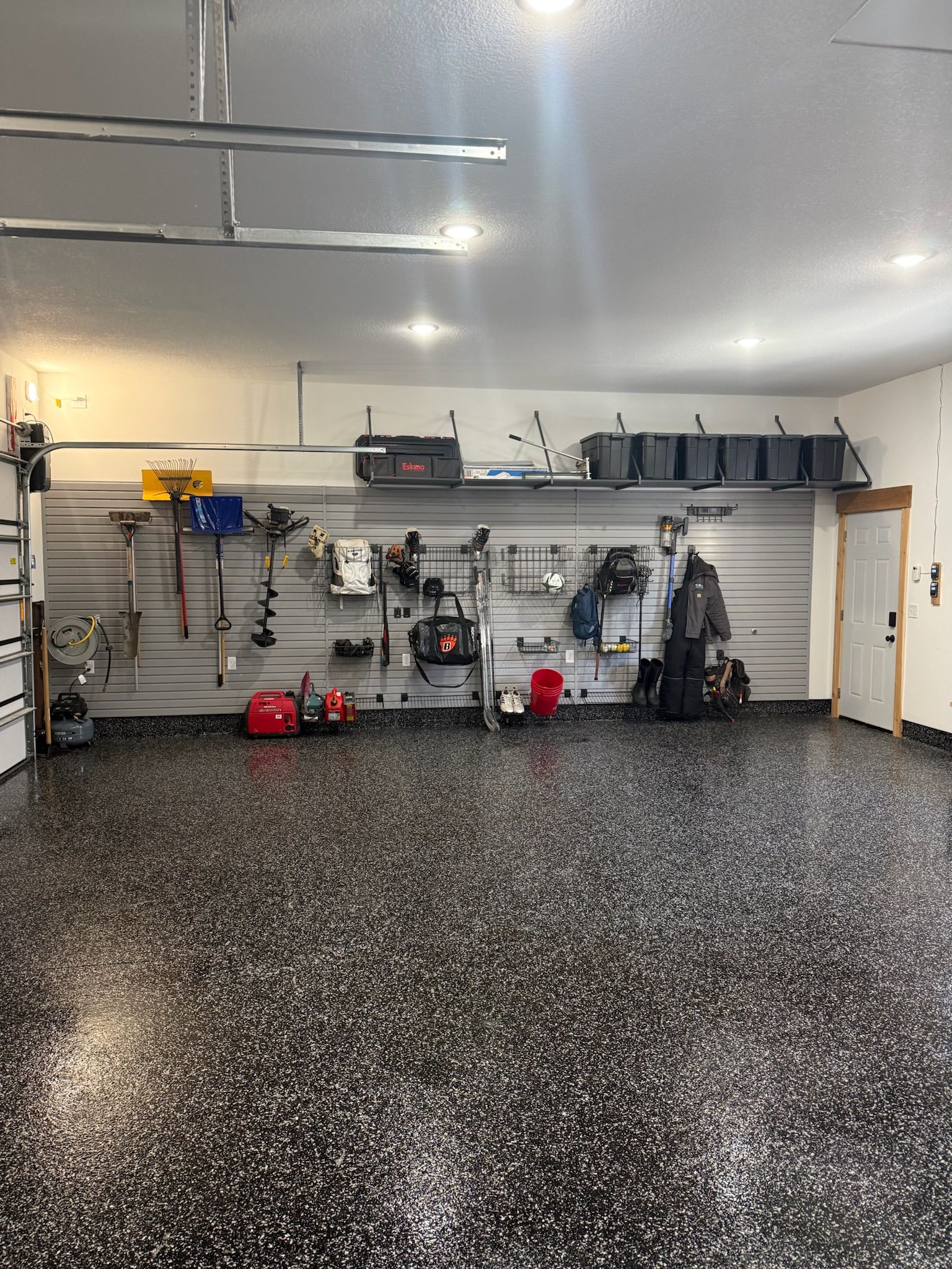 Garage interior with white walls, speckled floor, and organized tools, shelves, and equipment along the back wall