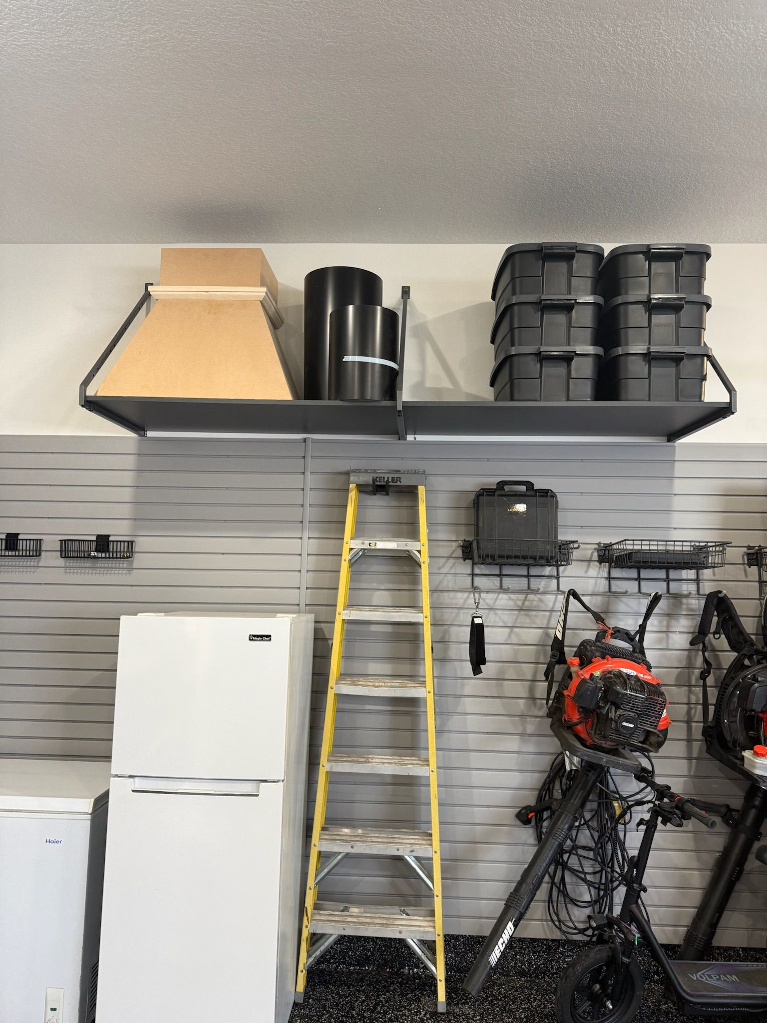 Garage storage shelves with bins, ladder, refrigerator, and hanging tools on a gray wall