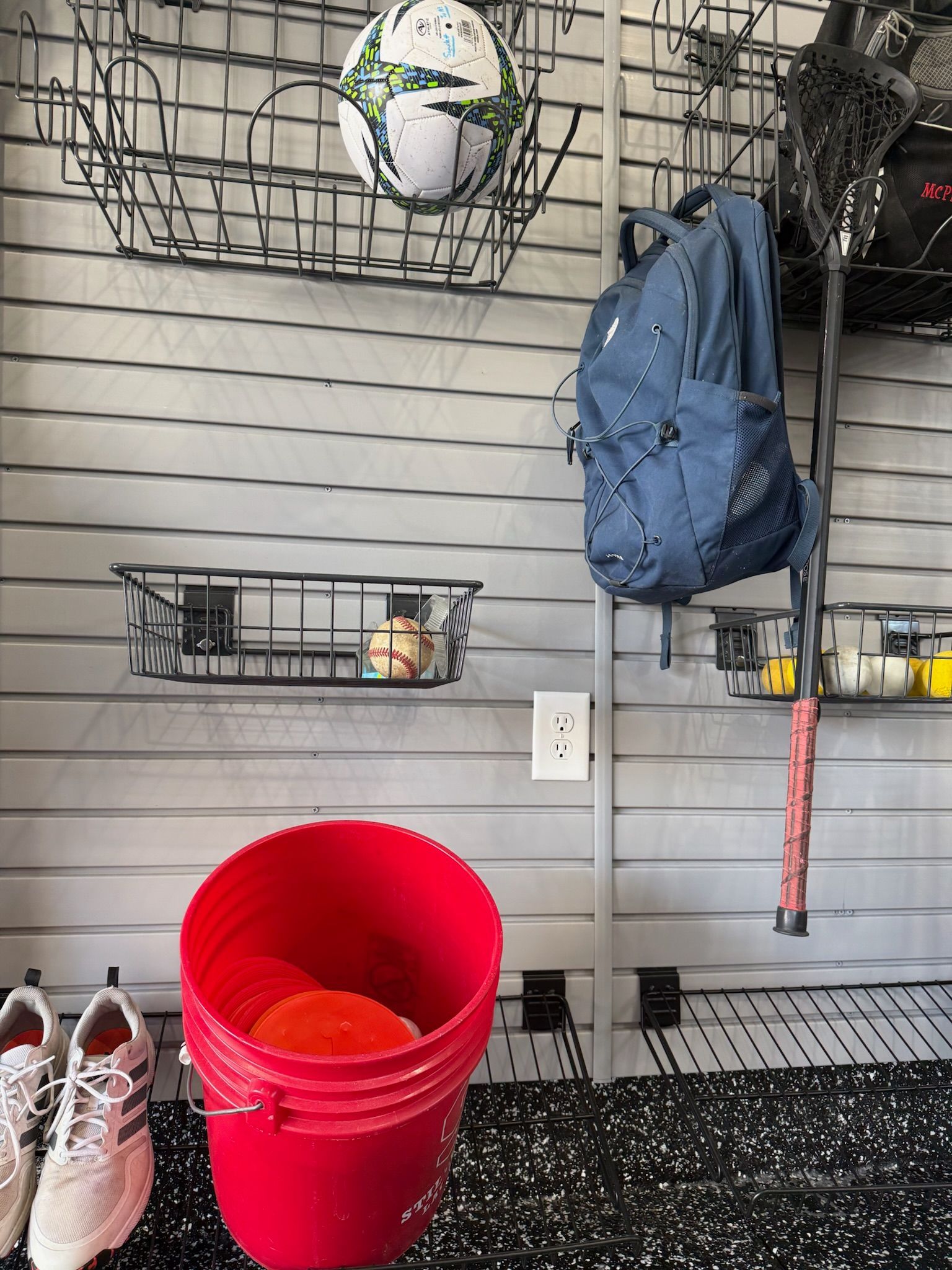 Garage wall with hanging blue backpack, shelf items, red bucket, and shoes on a speckled floor.