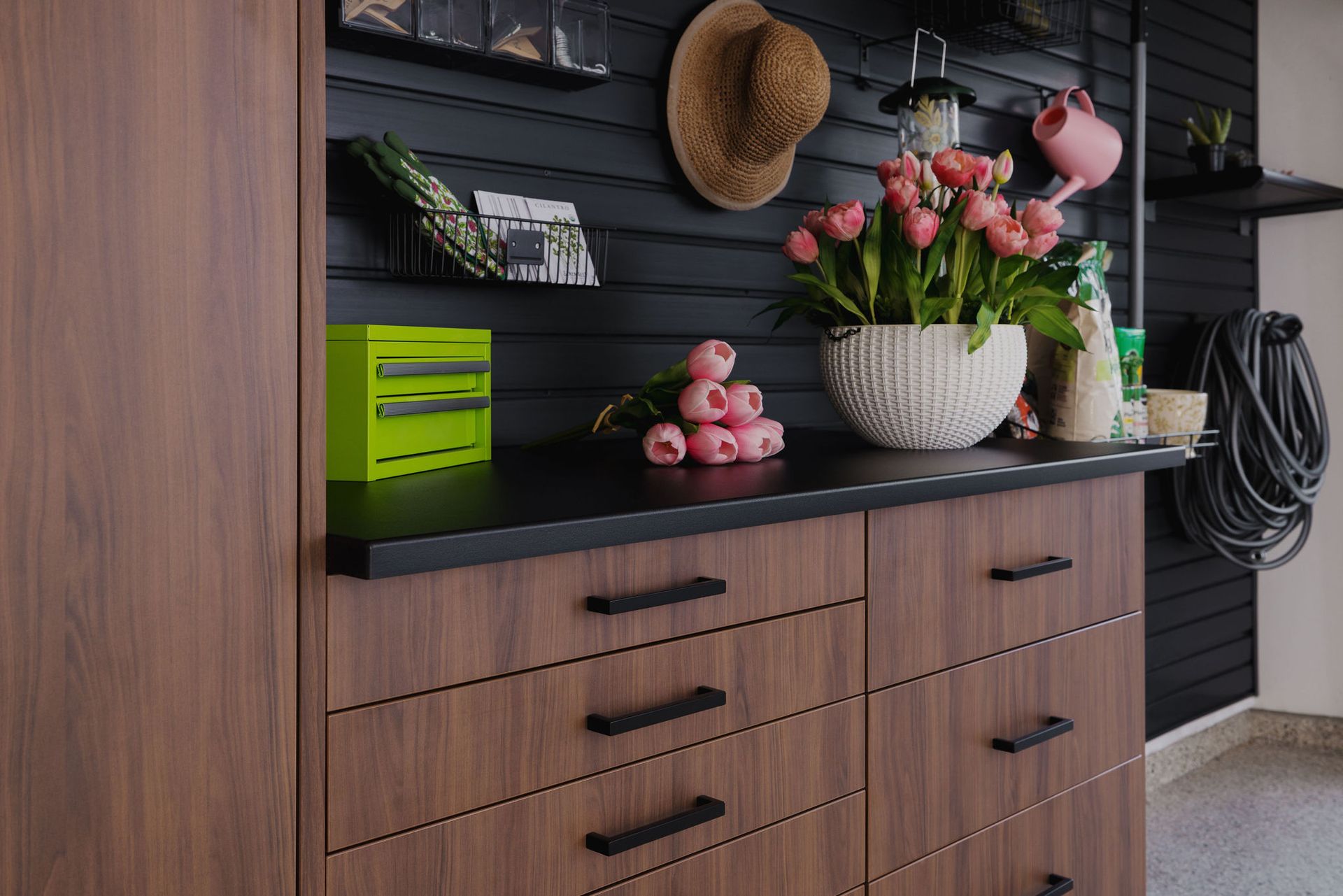 A wooden storage unit with drawers stands against a black slatted wall, featuring a potted plant, flowers, and a hat.