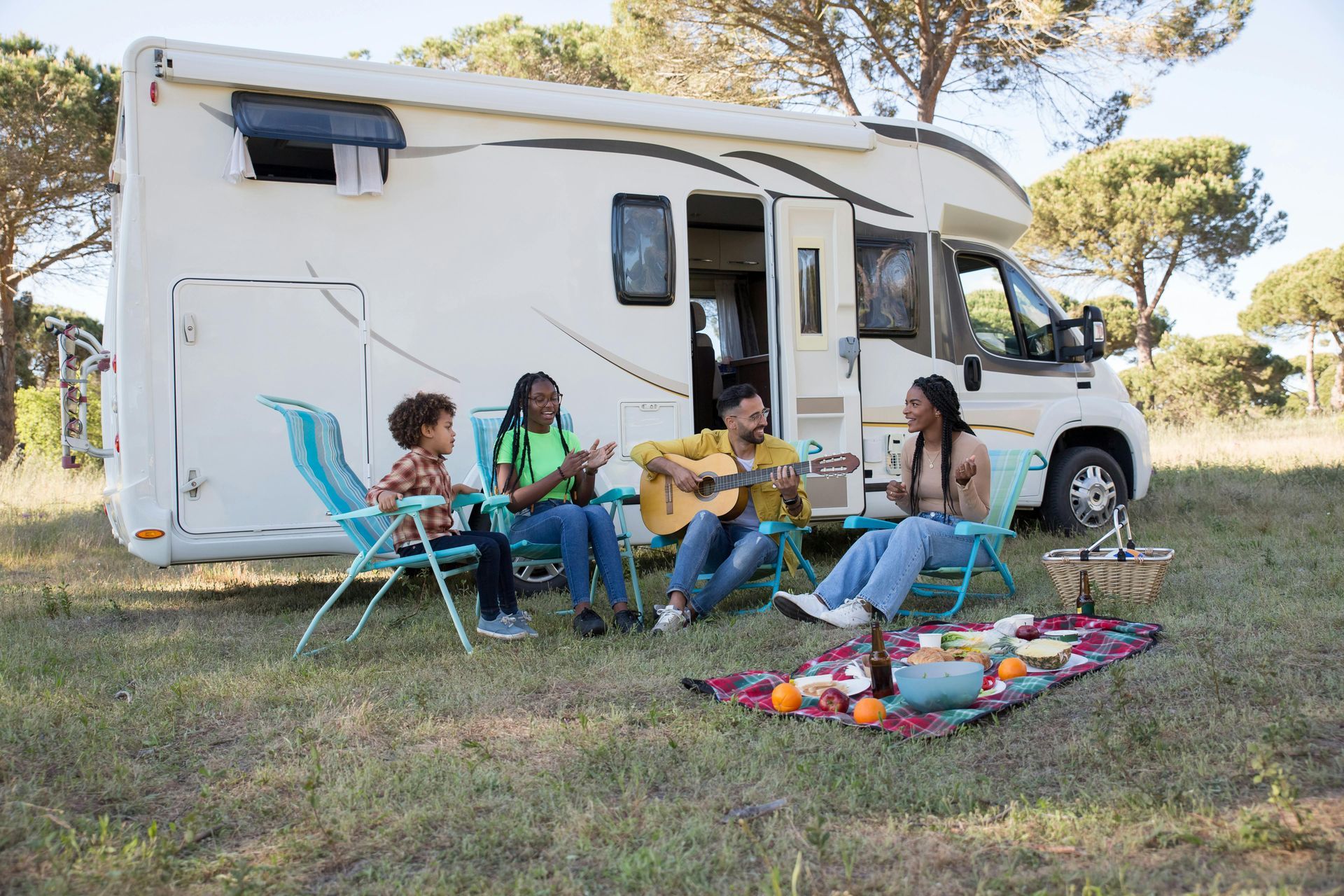Group sitting in camping chairs beside a white RV in a grassy field, sharing a picnic with colorful blankets.