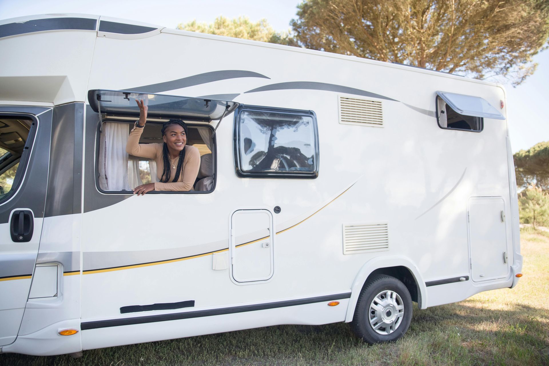 Woman leaning out of window of a white camper van parked on grass near trees