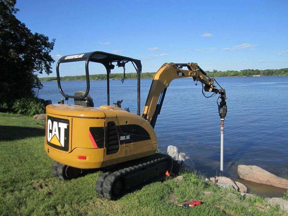 A cat excavator is sitting on the grass next to a body of water.