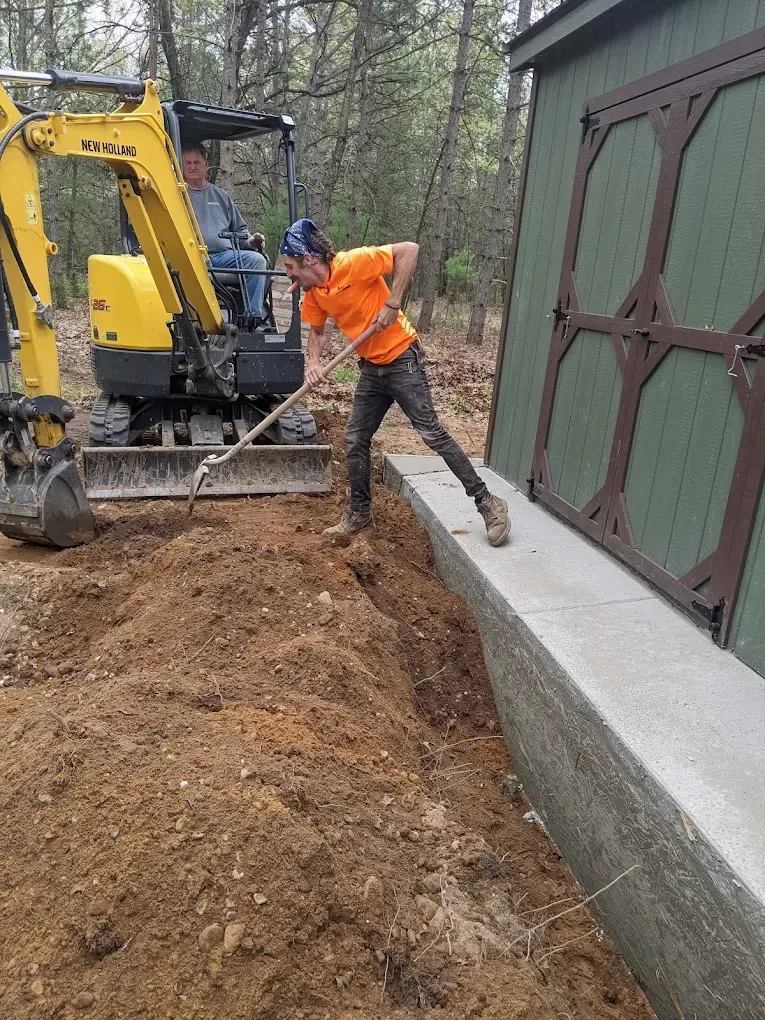 A man is digging in the dirt next to a yellow excavator.