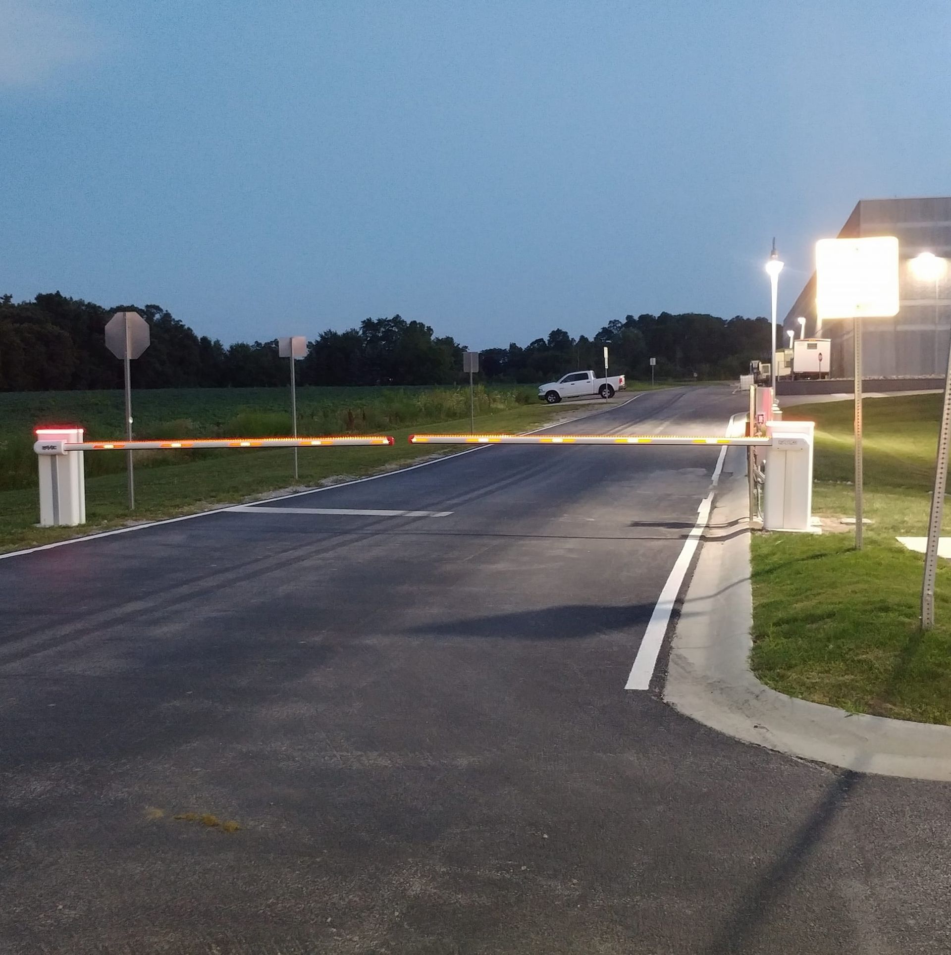 Empty paved entrance road at dusk with white gate posts and a lit building on the right.