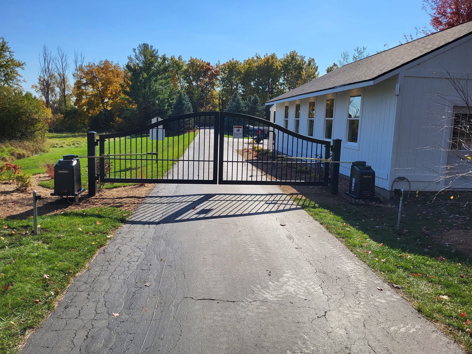 Driveway leading to a closed black gate beside a white house, with trees and lawn in the background