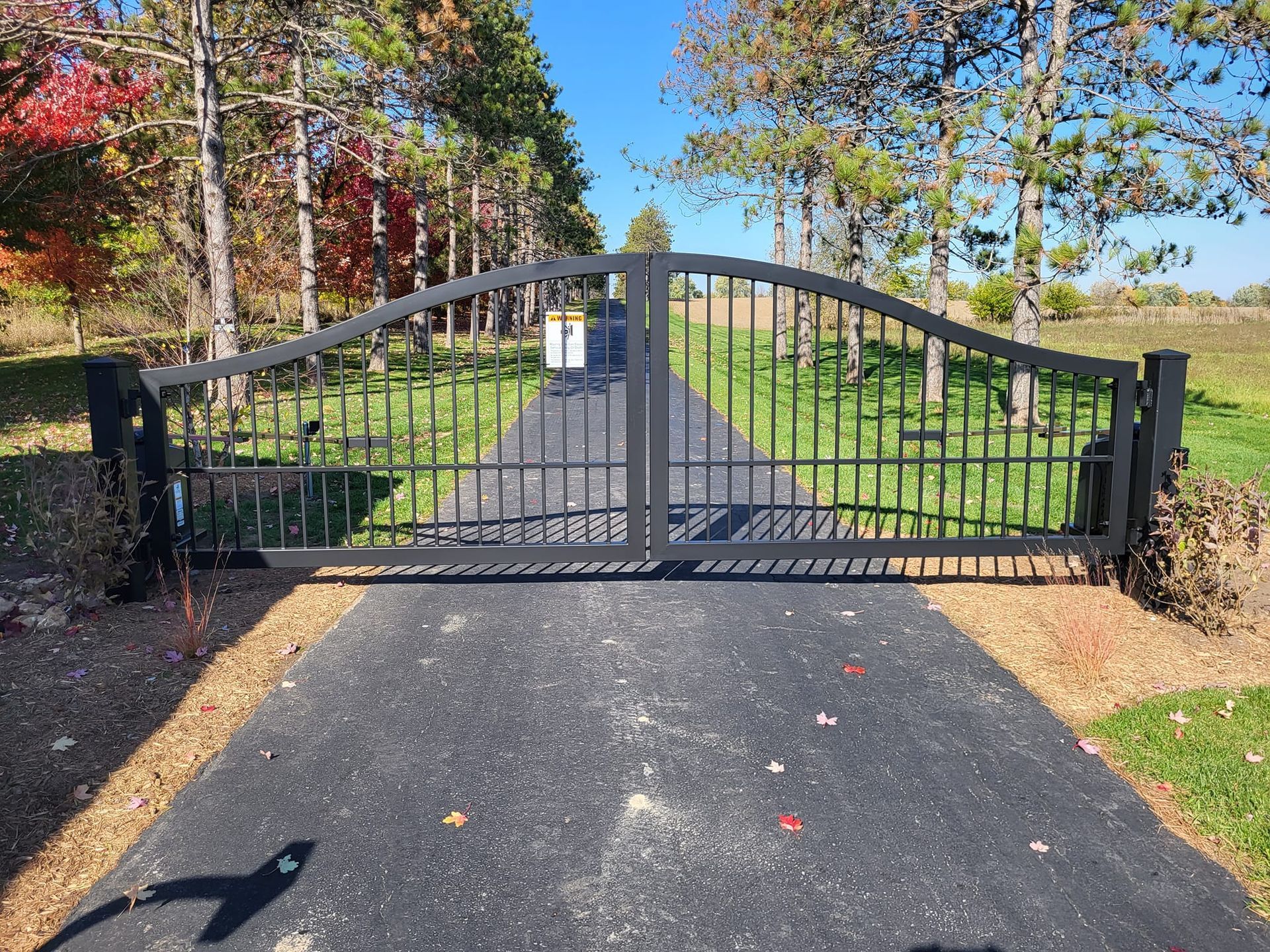 Black metal gate across a paved driveway with trees and grass beyond under a clear blue sky