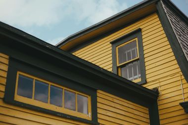 Yellow wooden house exterior with dark trim and windows against a cloudy sky