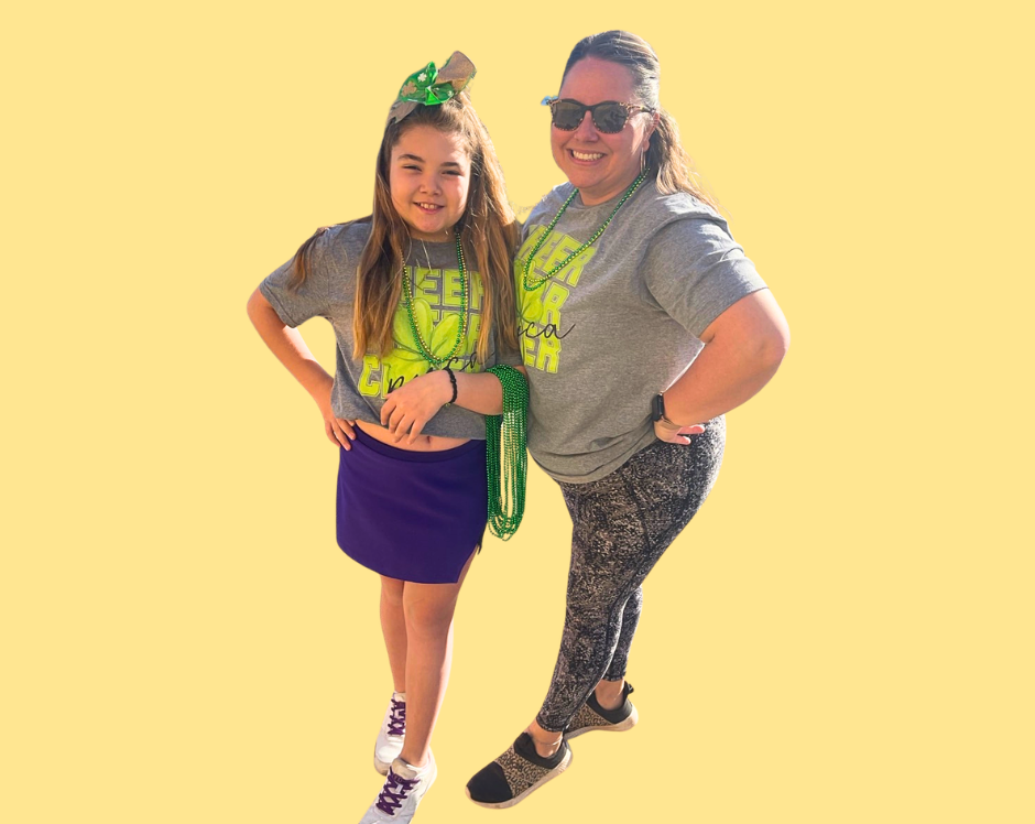 Football coach, arms crossed, on the field. Blue shirt, serious expression, players in background.
