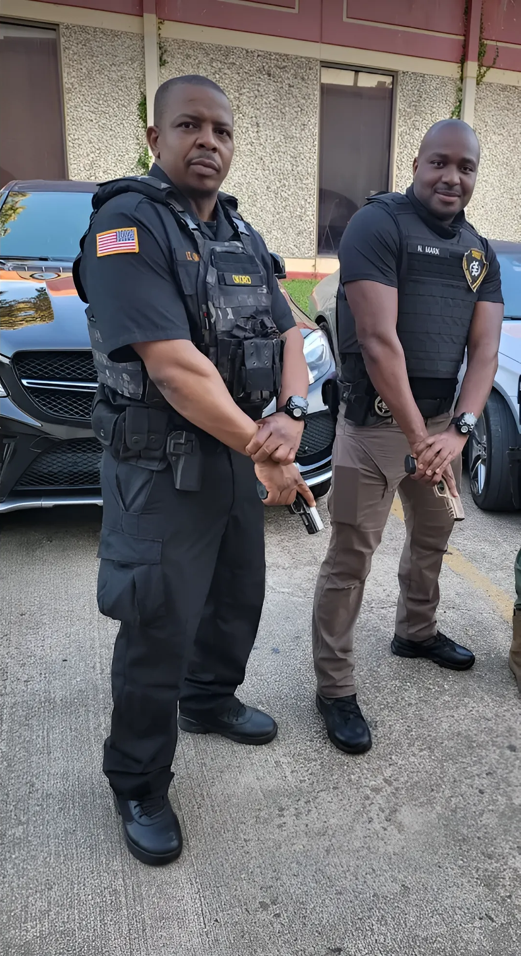 Two law enforcement officers standing outside. One wears a vest, the other wears a t-shirt. Both have their hands clasped.